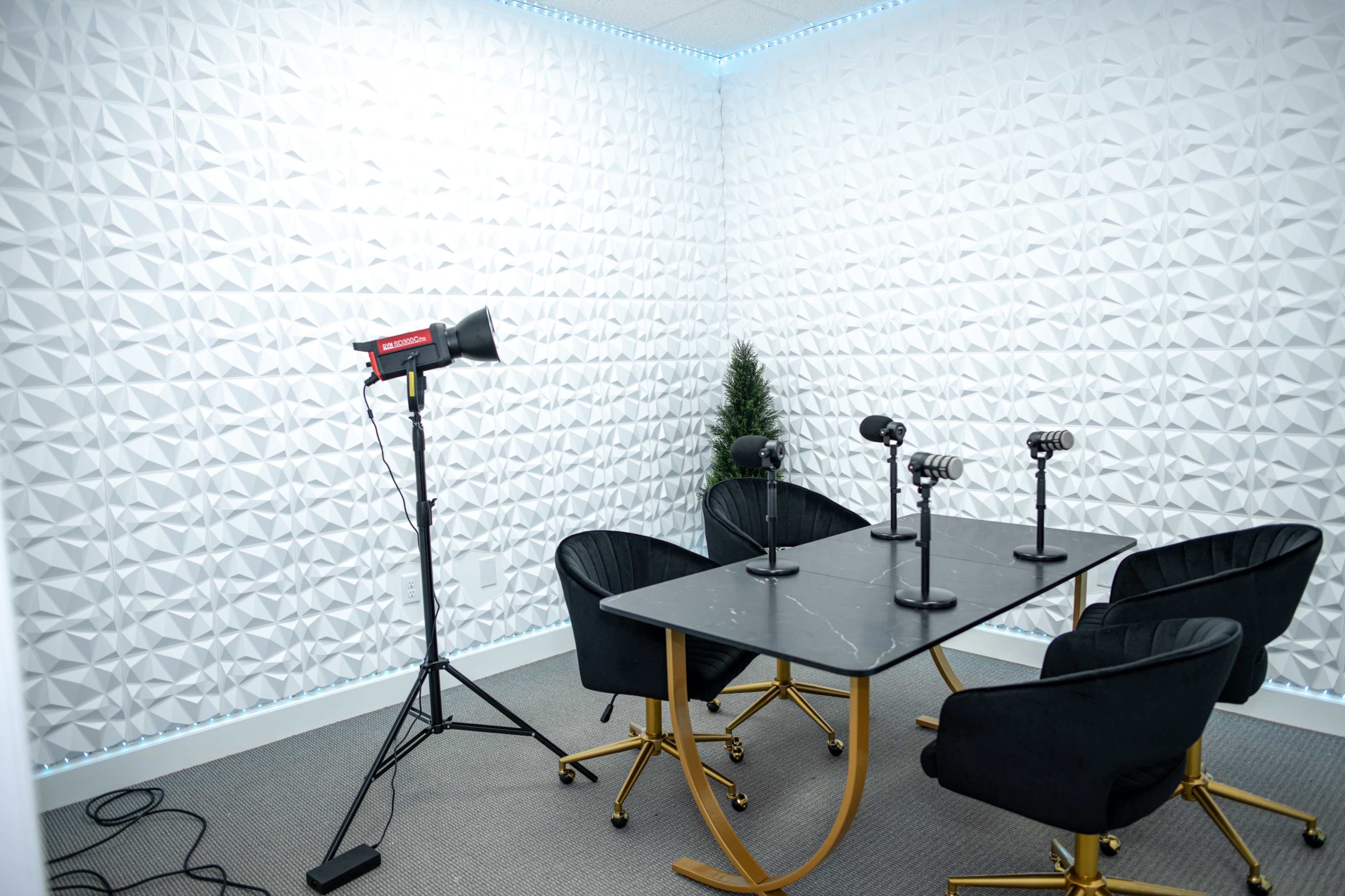 A small room features a table with four microphones and black chairs, set against a textured white wall and illuminated by soft lighting.