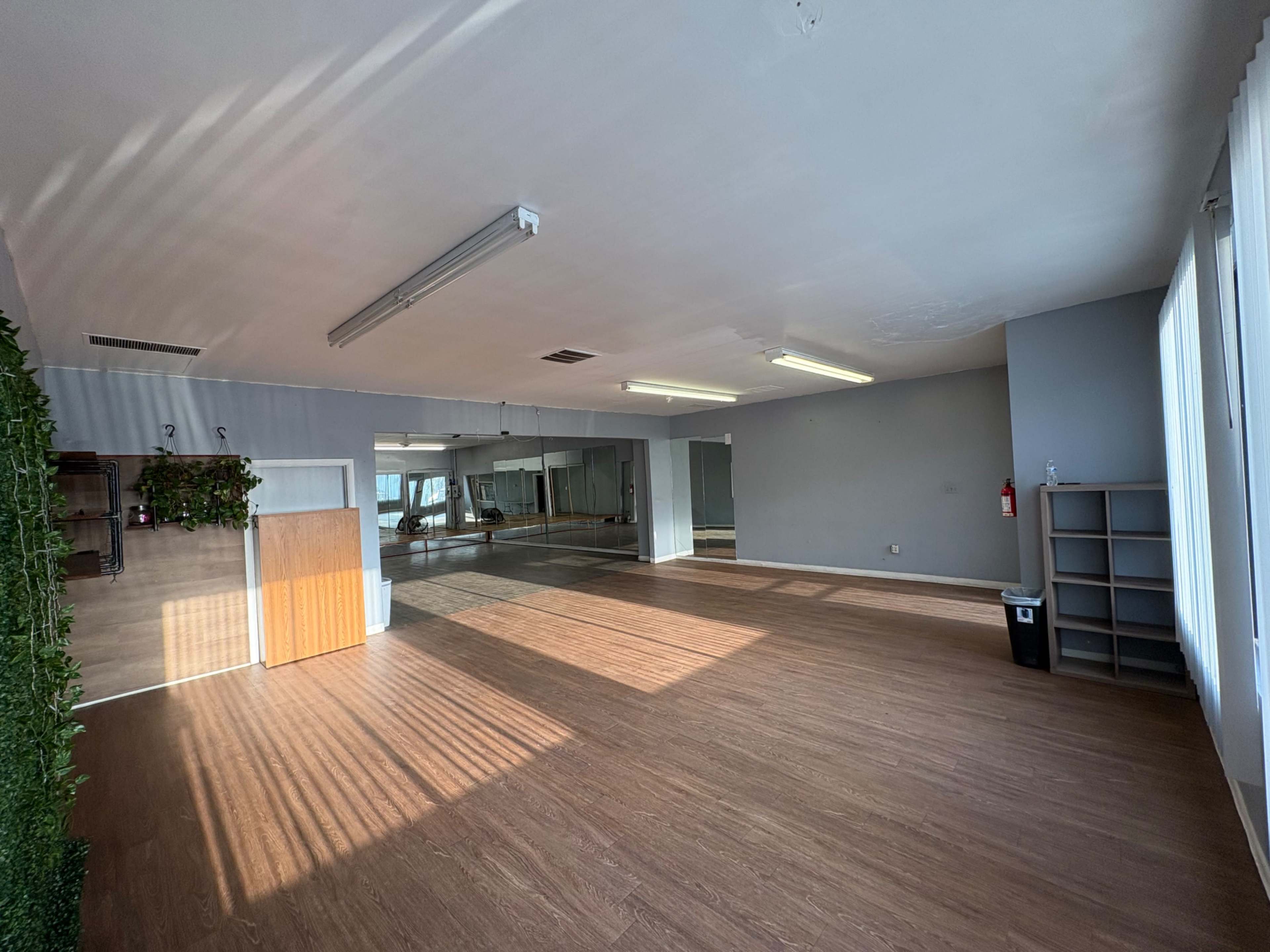 The image shows a spacious, well-lit room with wooden flooring, large windows, and mirrored walls, featuring a storage shelf and a door on one side.