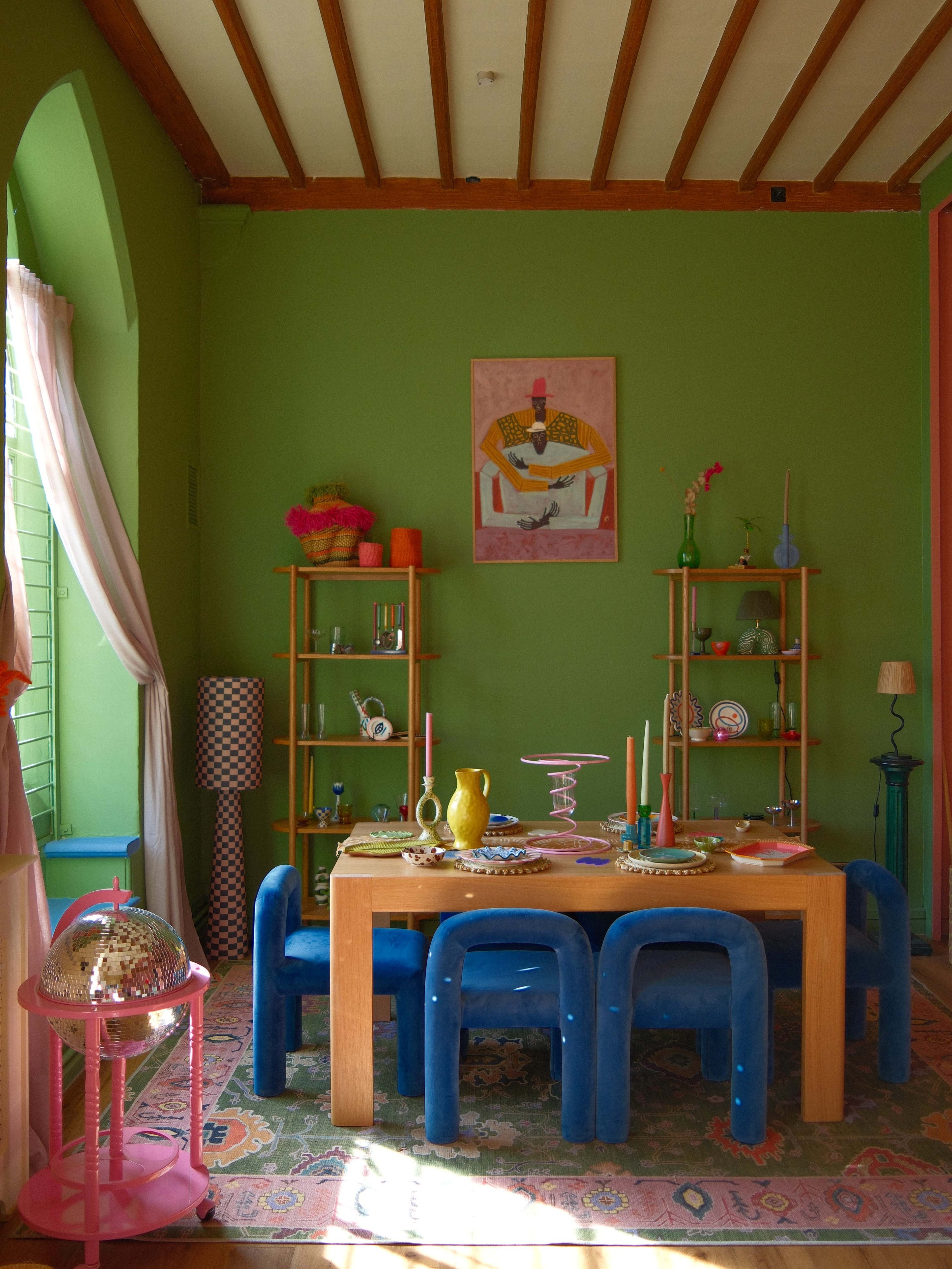 The image shows a brightly colored dining area with a wooden table surrounded by blue chairs, decorative shelves, and various tabletop items against a green wall.