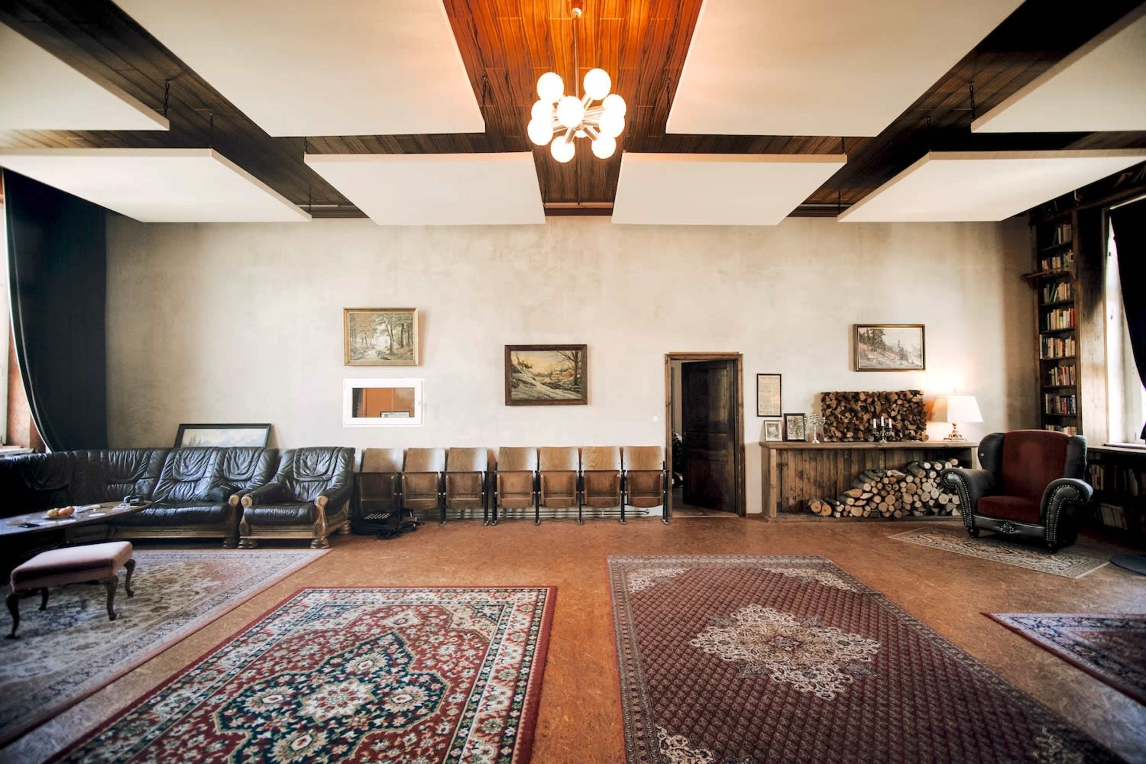 The image shows a spacious room with a leather sofa, a row of chairs, a rug, and paintings on the walls, illuminated by a overhead light fixture.