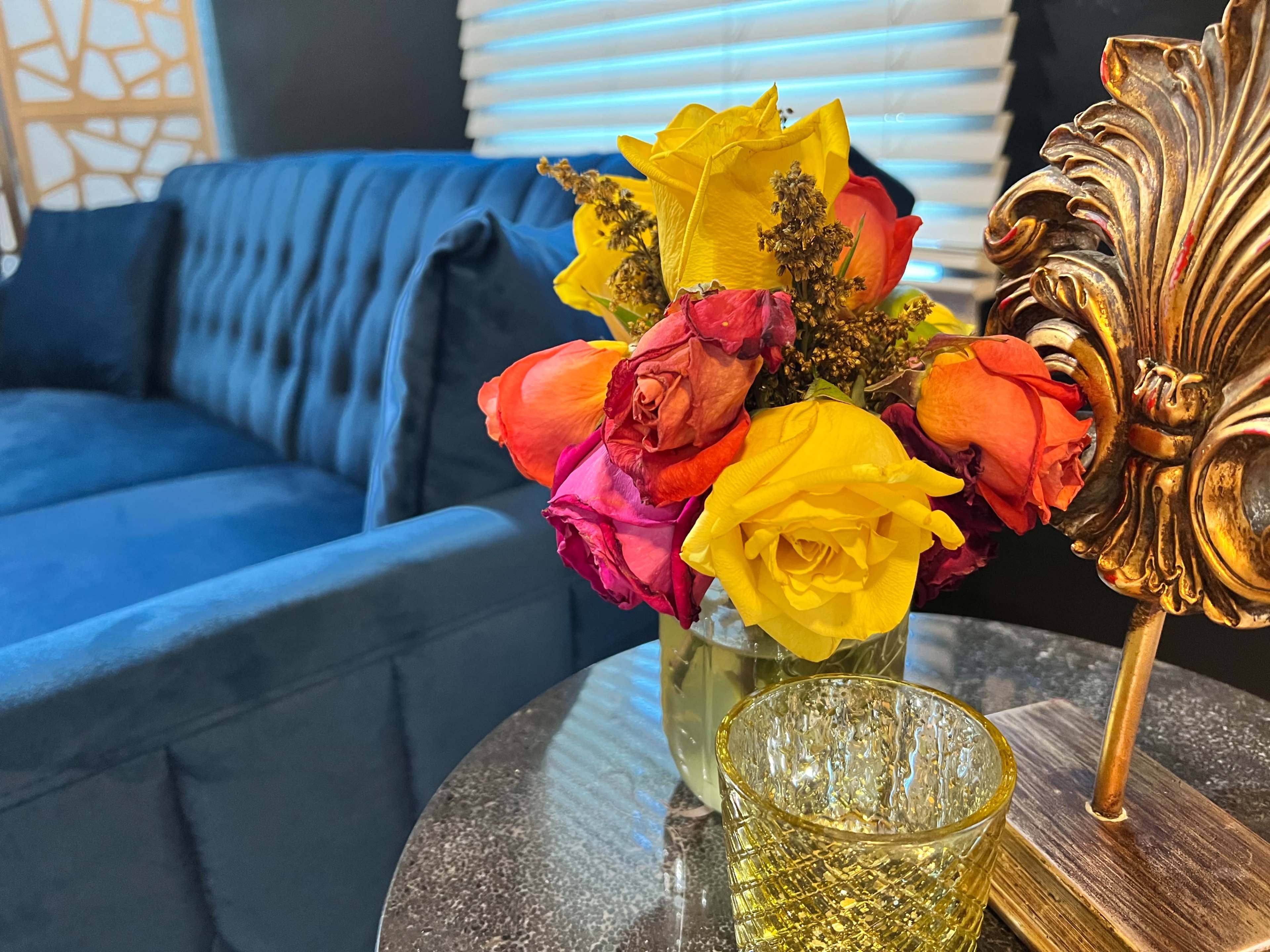 A bouquet of multicolored roses sits on a glass table next to a decorative gold item, with a blue sofa in the background.