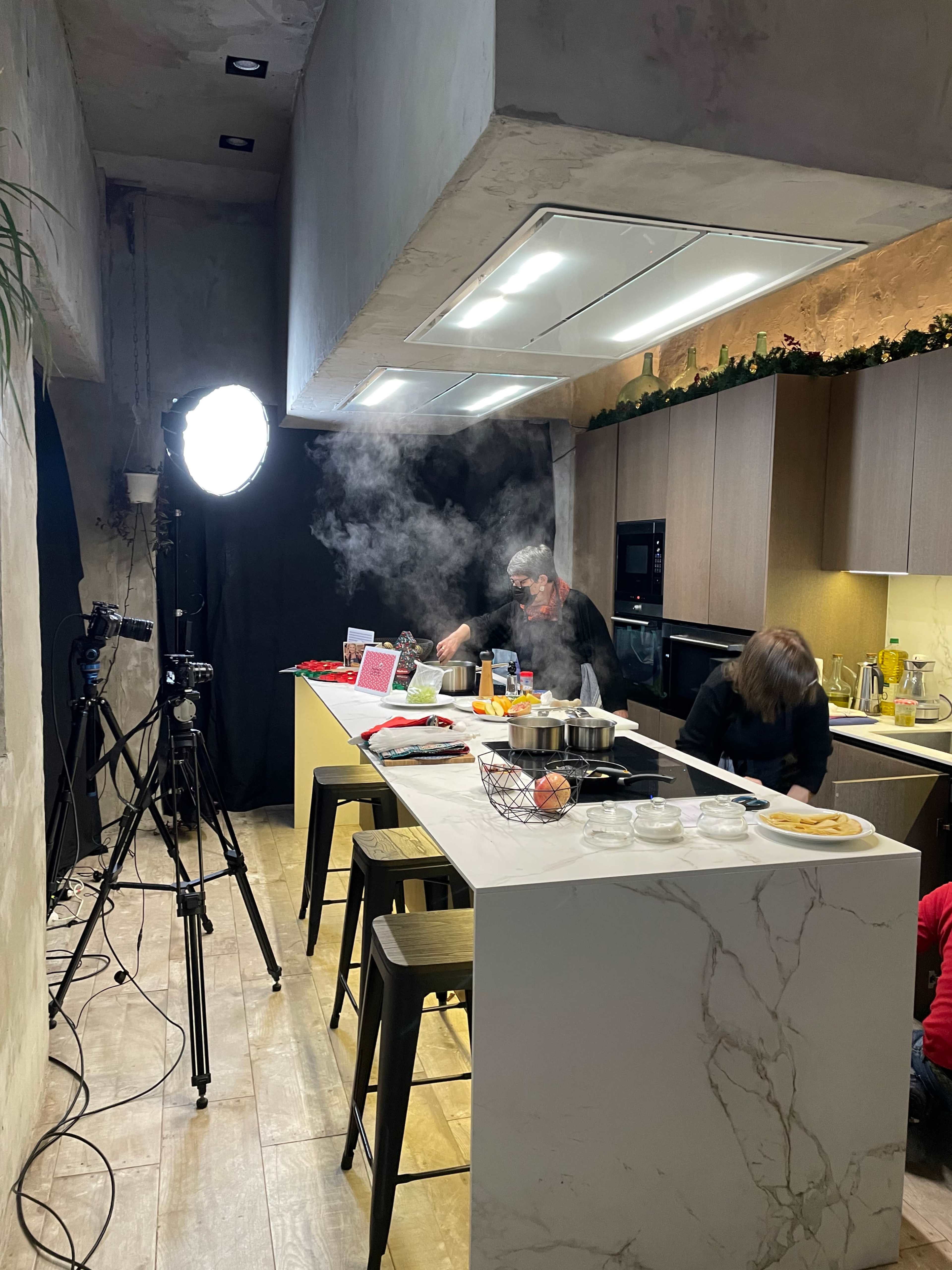 A kitchen scene features two people cooking at a countertop surrounded by cameras and lighting equipment, with steam rising from pots on the stove.