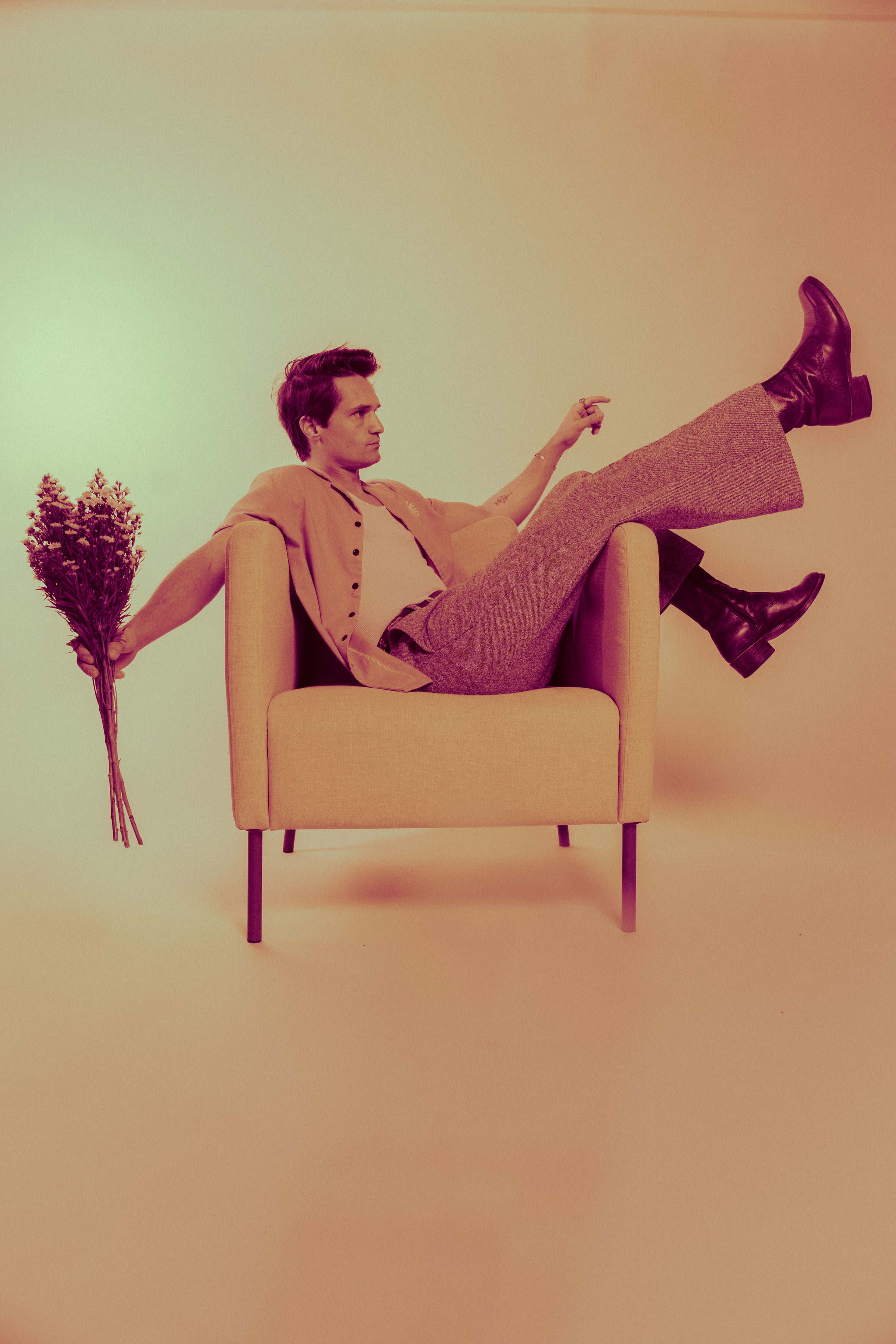 A person in a light-colored outfit poses casually in a chair while holding a bouquet of flowers.