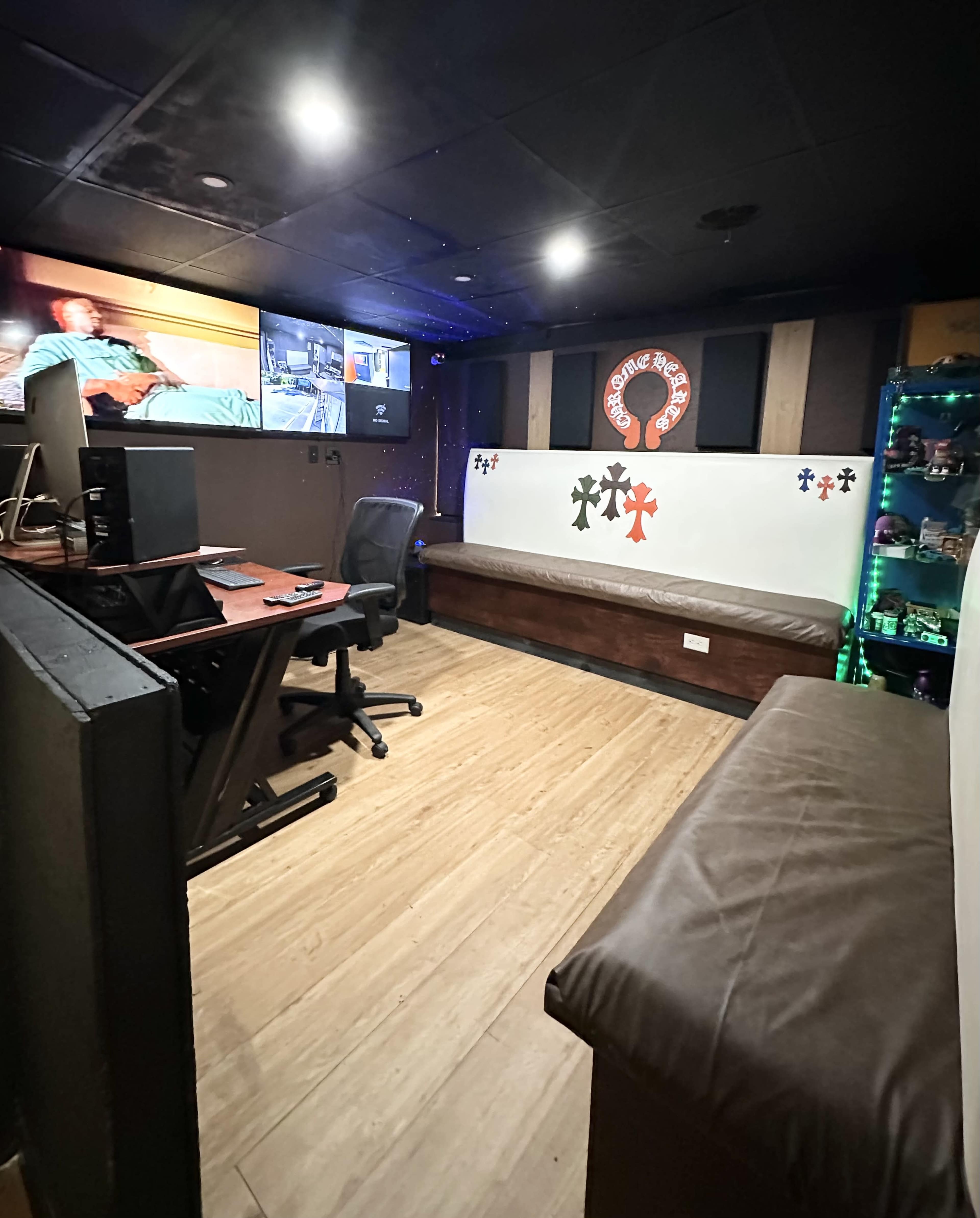 The image shows a small room featuring a desk with a computer, a seating area with a brown bench, and wall-mounted monitors displaying video feeds.