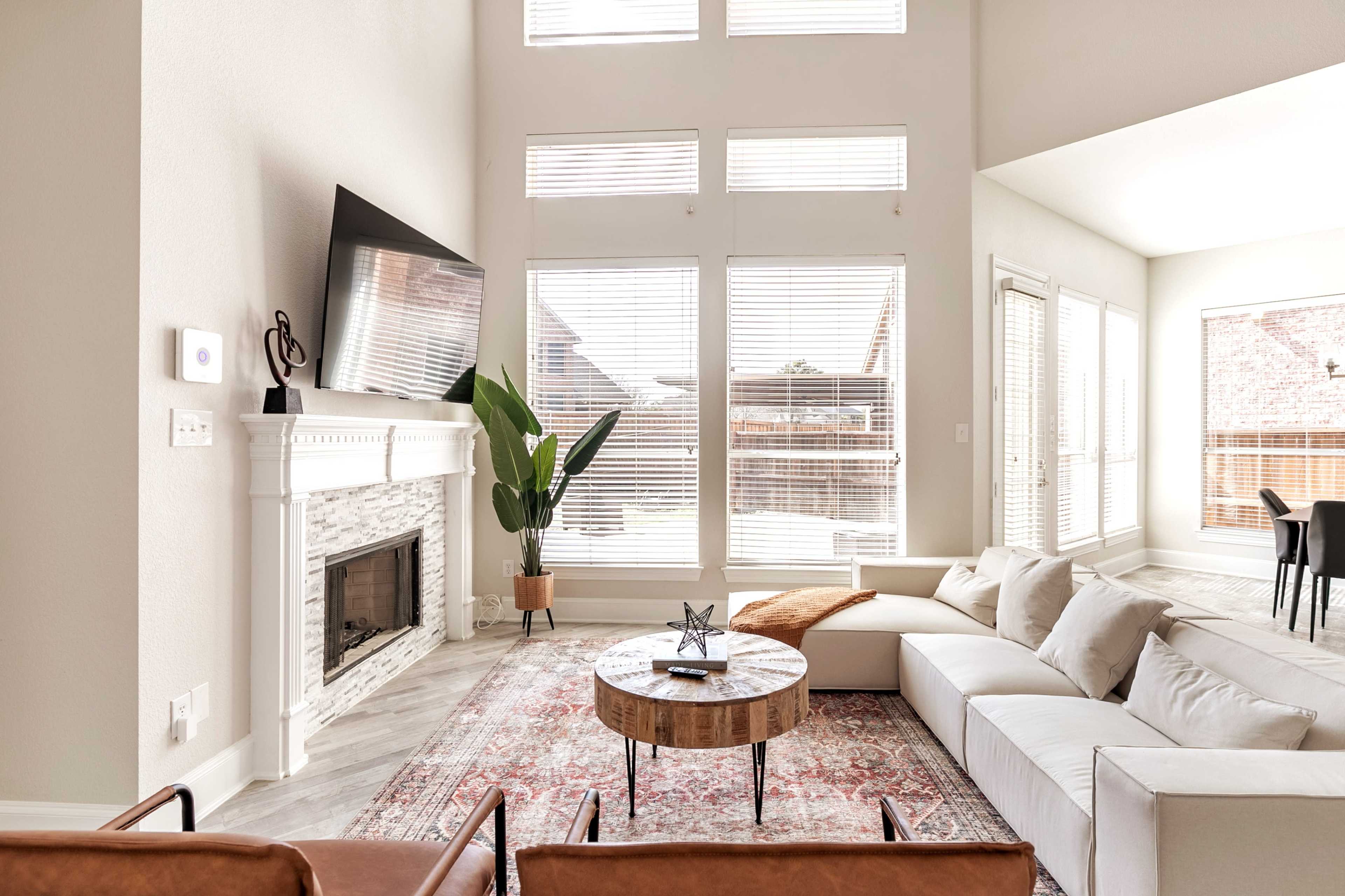 A spacious living room features a large sectional sofa, a circular coffee table, a fireplace, and tall windows allowing natural light to enter.