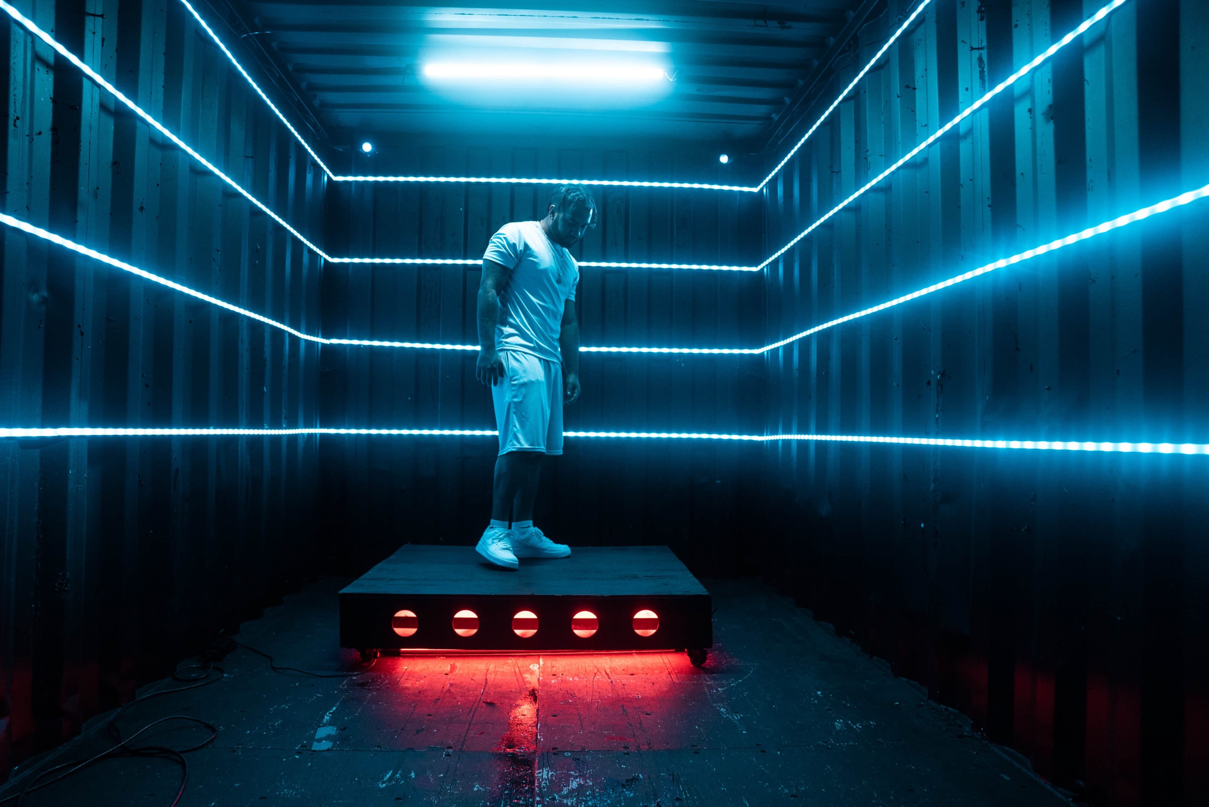 A person stands on a raised platform inside a shipping container with blue and red neon lights illuminating the walls.