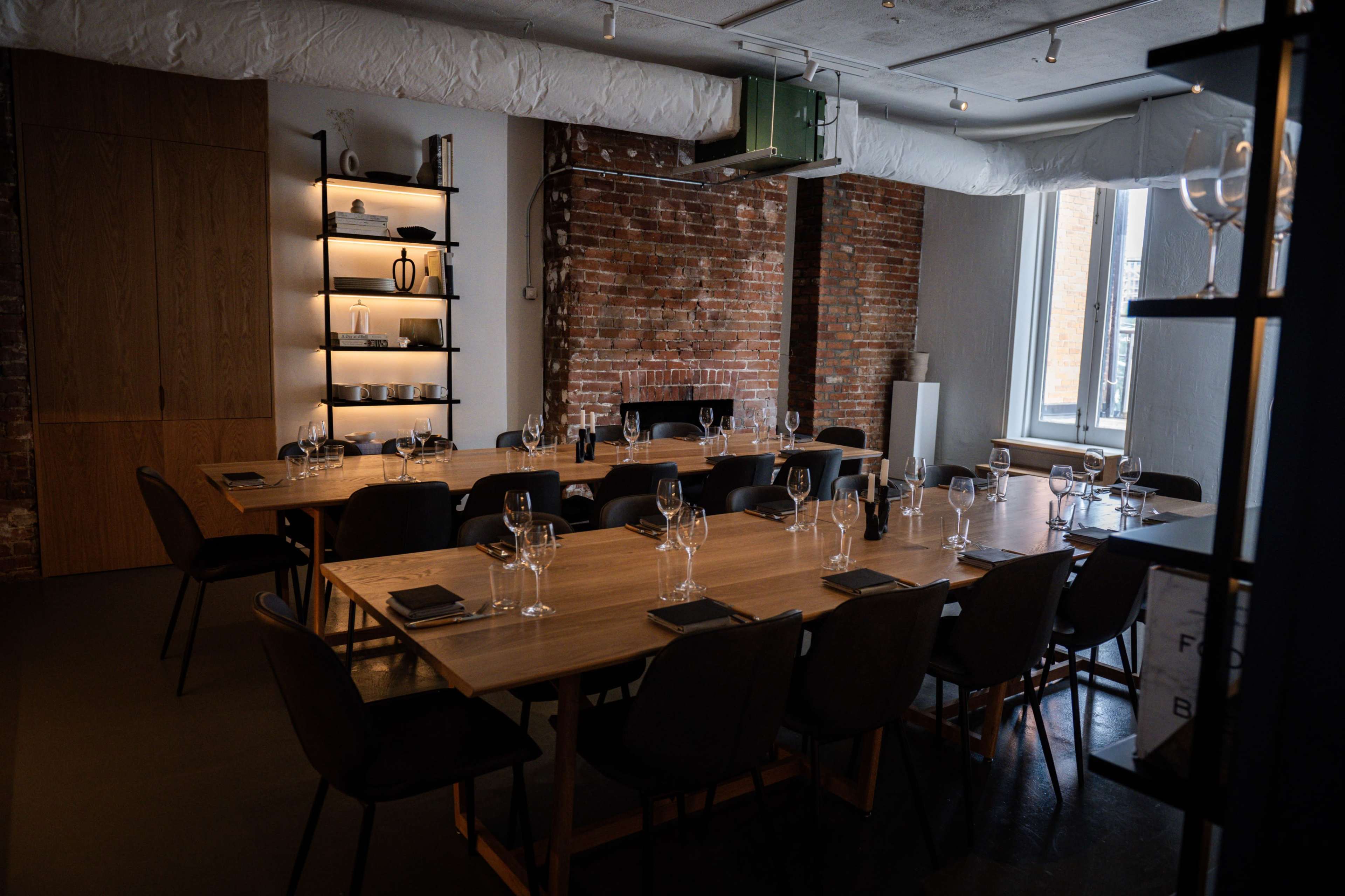 A dining room features a long wooden table set with glasses and cutlery, surrounded by dark chairs against a backdrop of exposed brick walls and industrial decor.