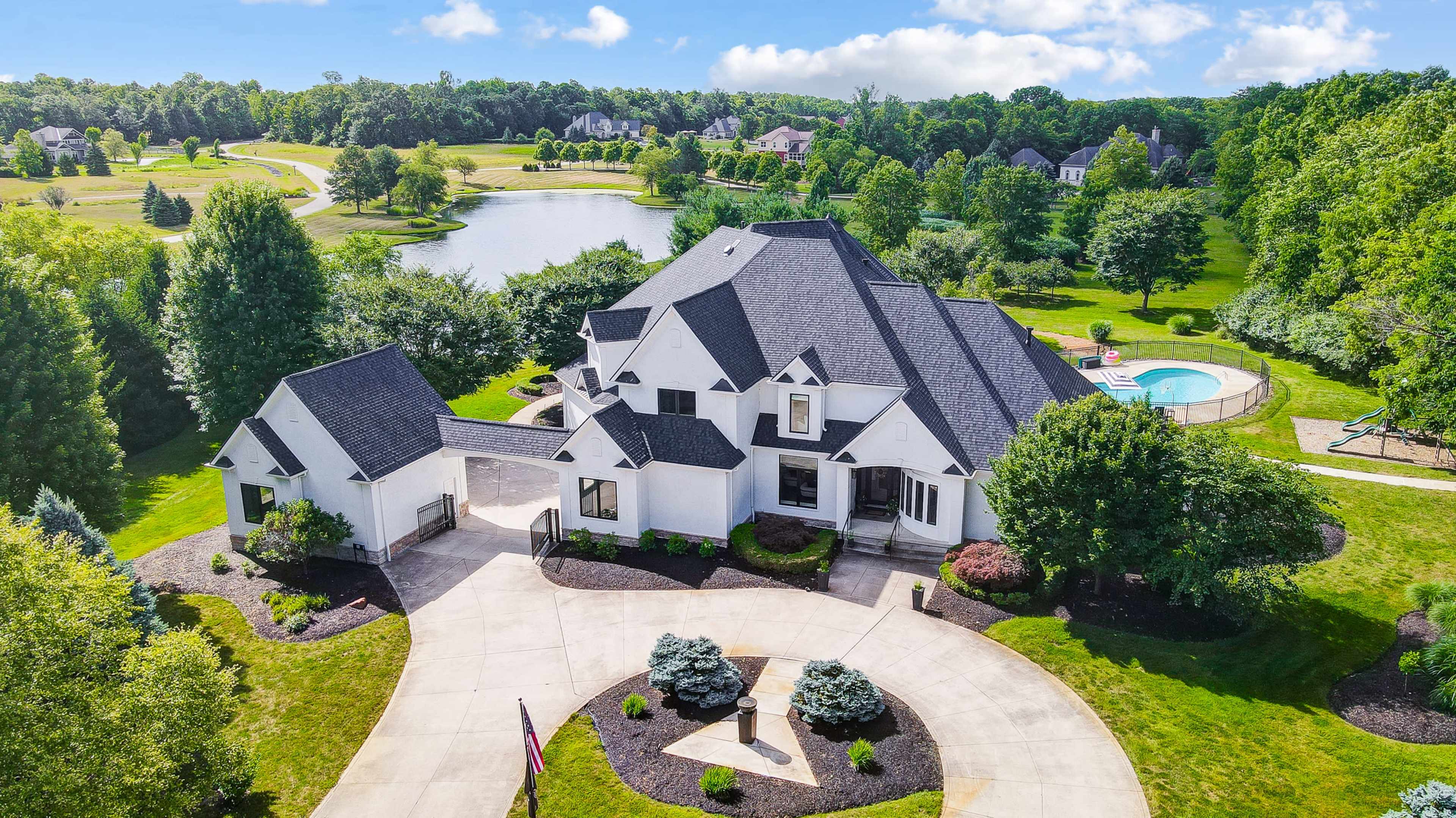 A large, modern home with a prominent roof design is situated on a landscaped property near a pond and swimming pool.