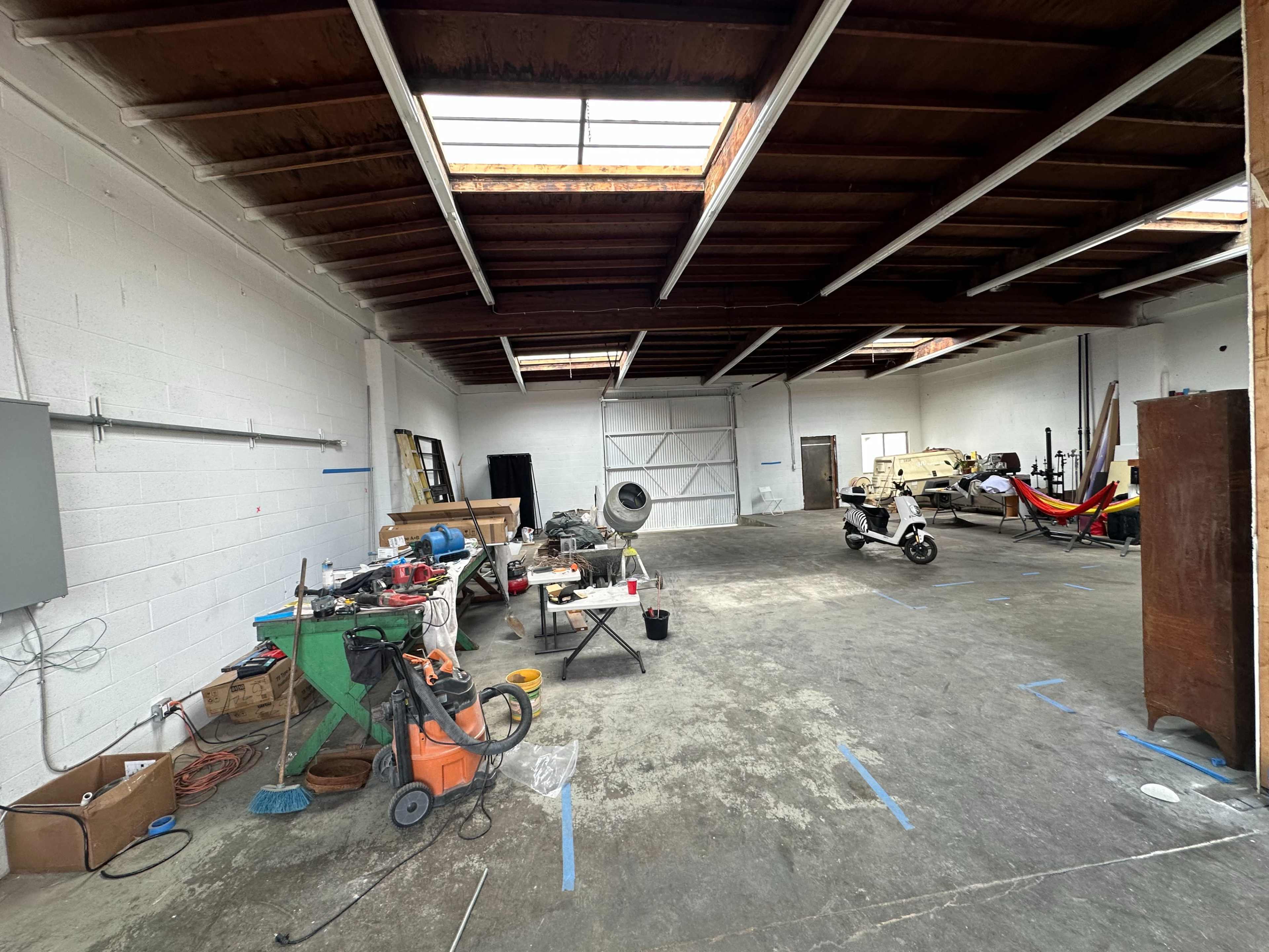 The image shows a spacious, empty warehouse with high ceilings, stacked with various tools, equipment, and furniture.