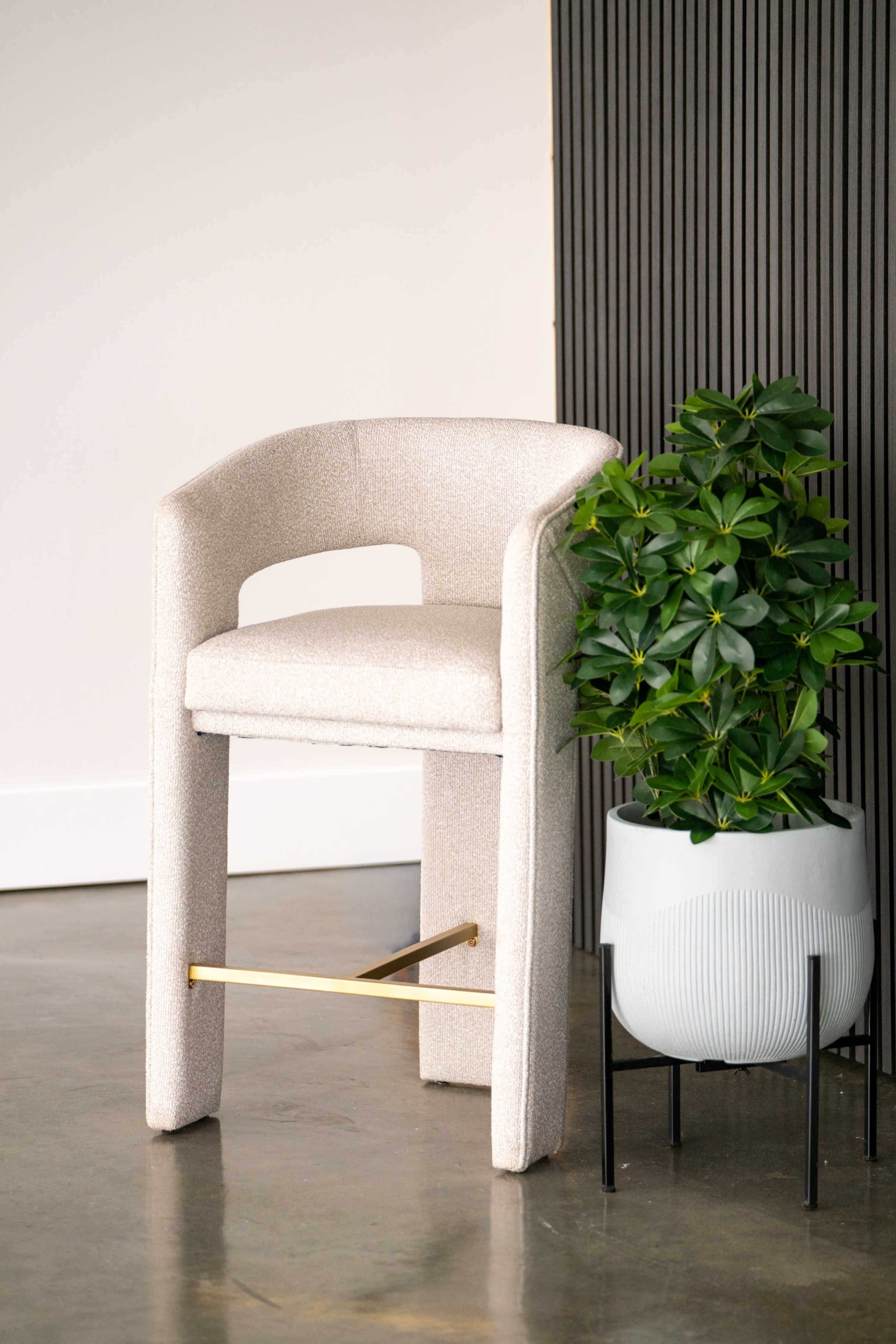 A beige upholstered stool is positioned next to a potted plant in a modern planter against a textured wall.
