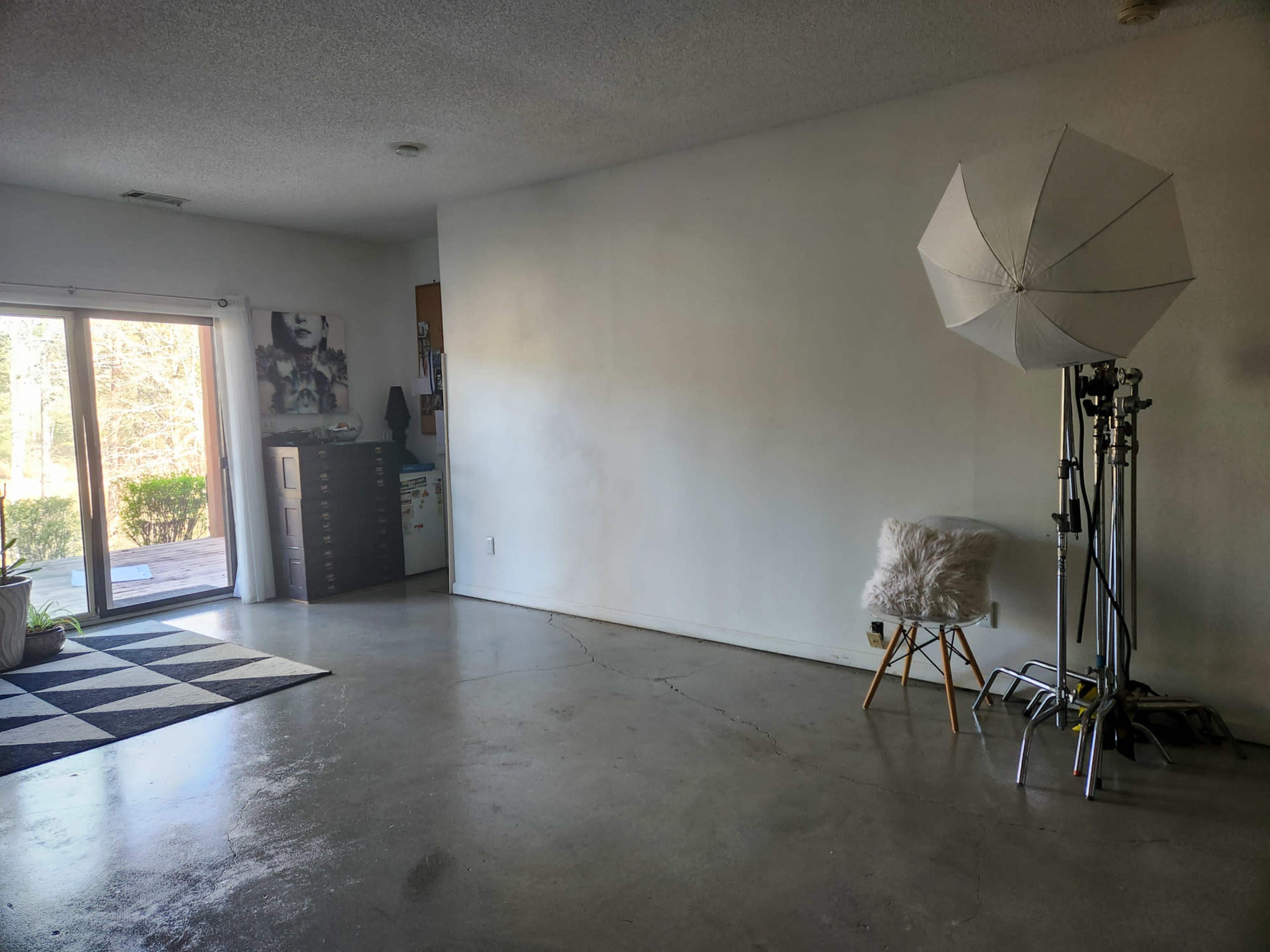 The image shows a spacious, empty room with a white wall, a photography umbrella stand, a chair with a faux fur throw, and a checkered rug in one corner.