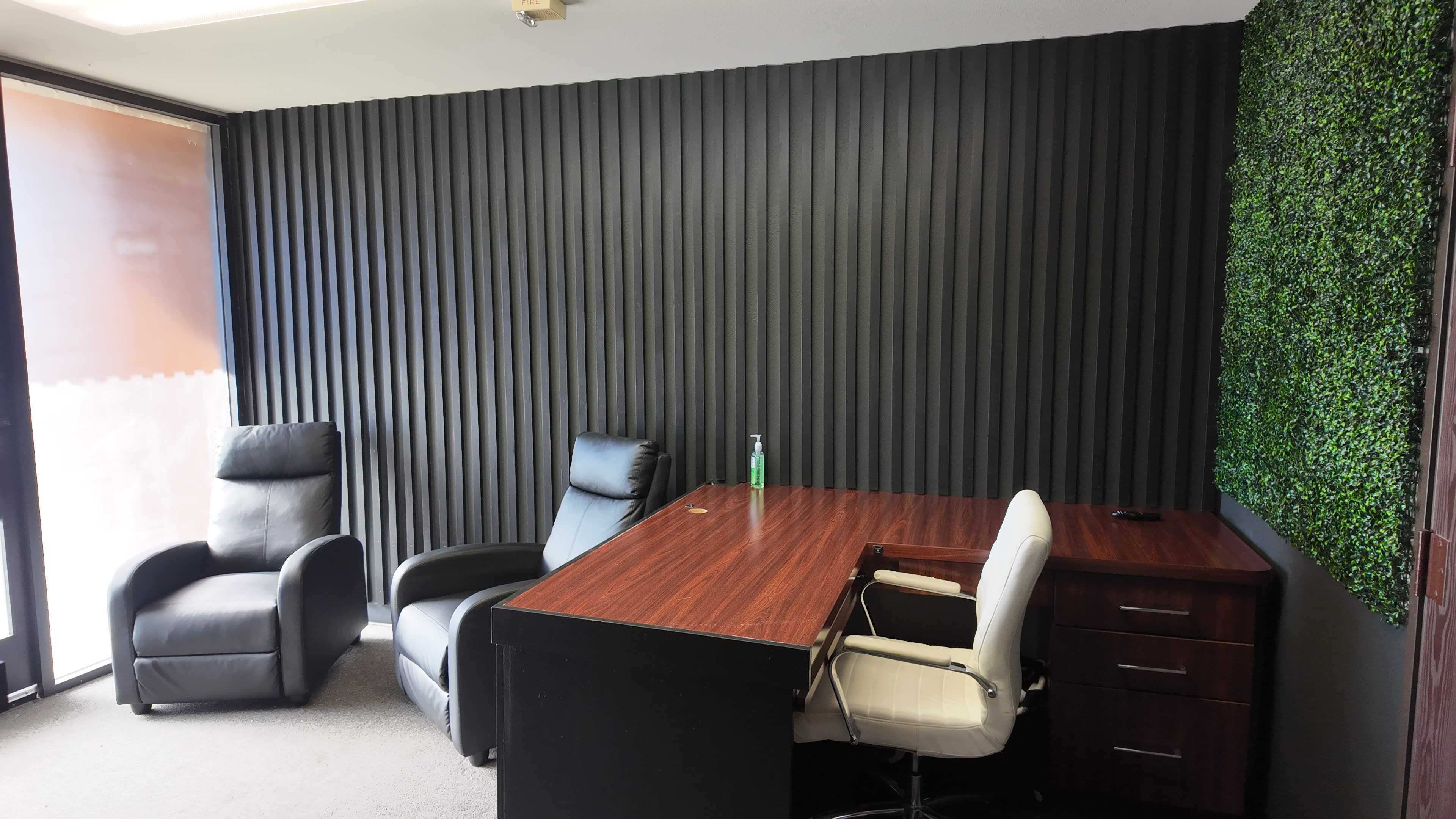The image shows a sparsely furnished office space with a dark wall featuring vertical stripes, a wooden desk, two chairs—one white and one dark leather—and a section with artificial greenery.