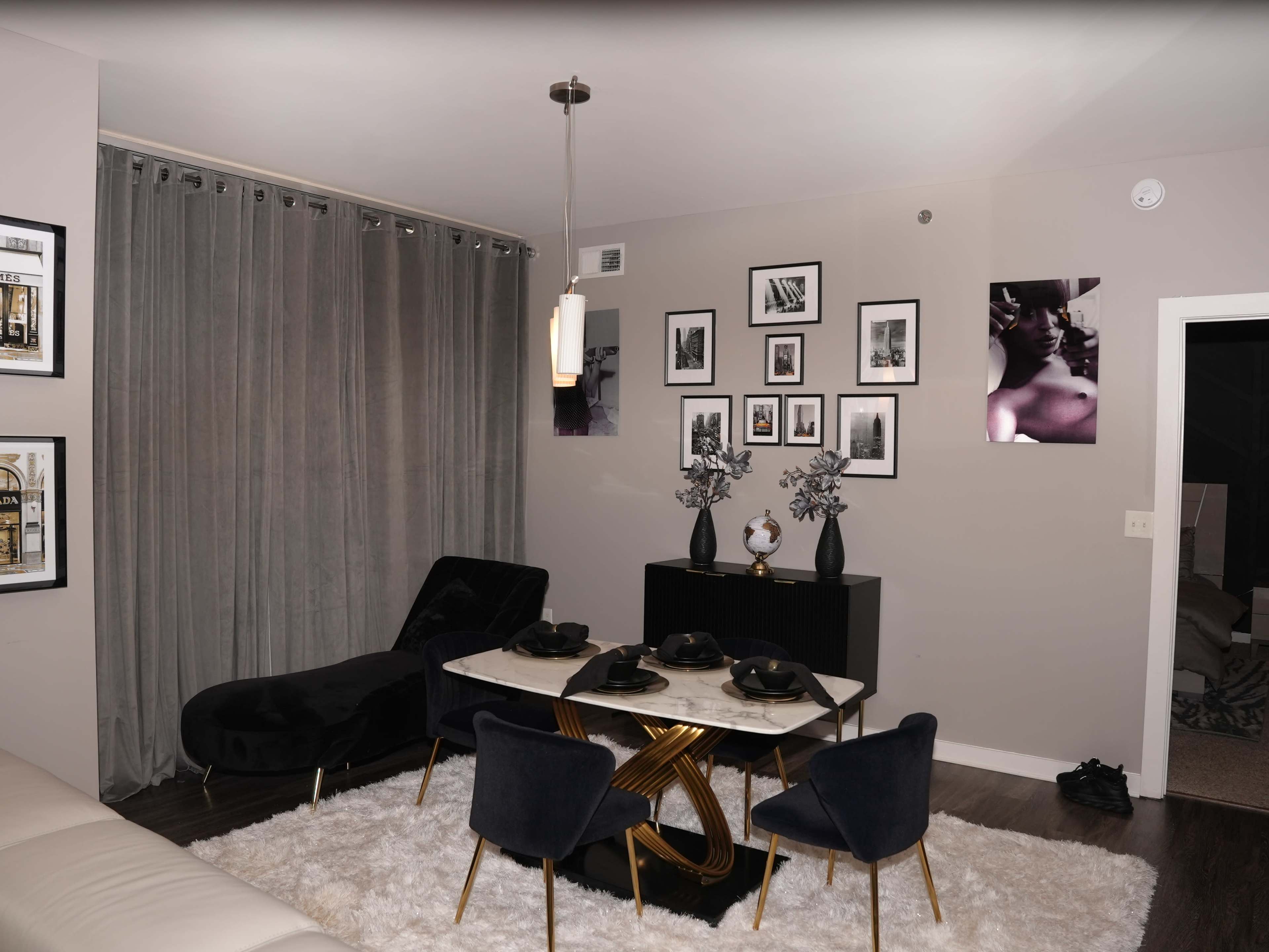 The scene features a modern dining area with a marble table and four black chairs, surrounded by gray curtains and framed photographs on the walls.