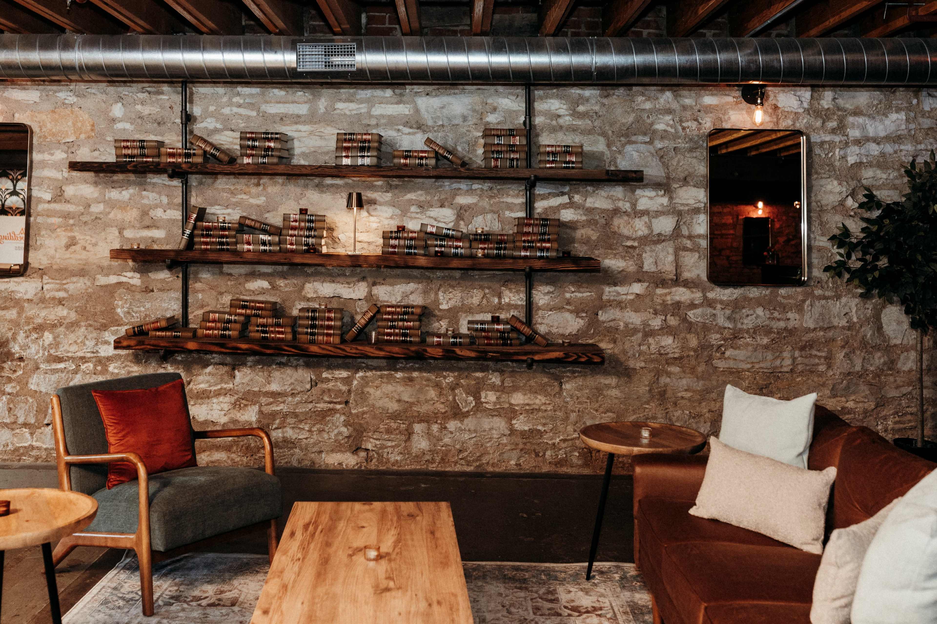 The image shows a cozy interior with a stone wall, featuring wooden shelves filled with books, a brown sofa, and a small round table with metal accents.