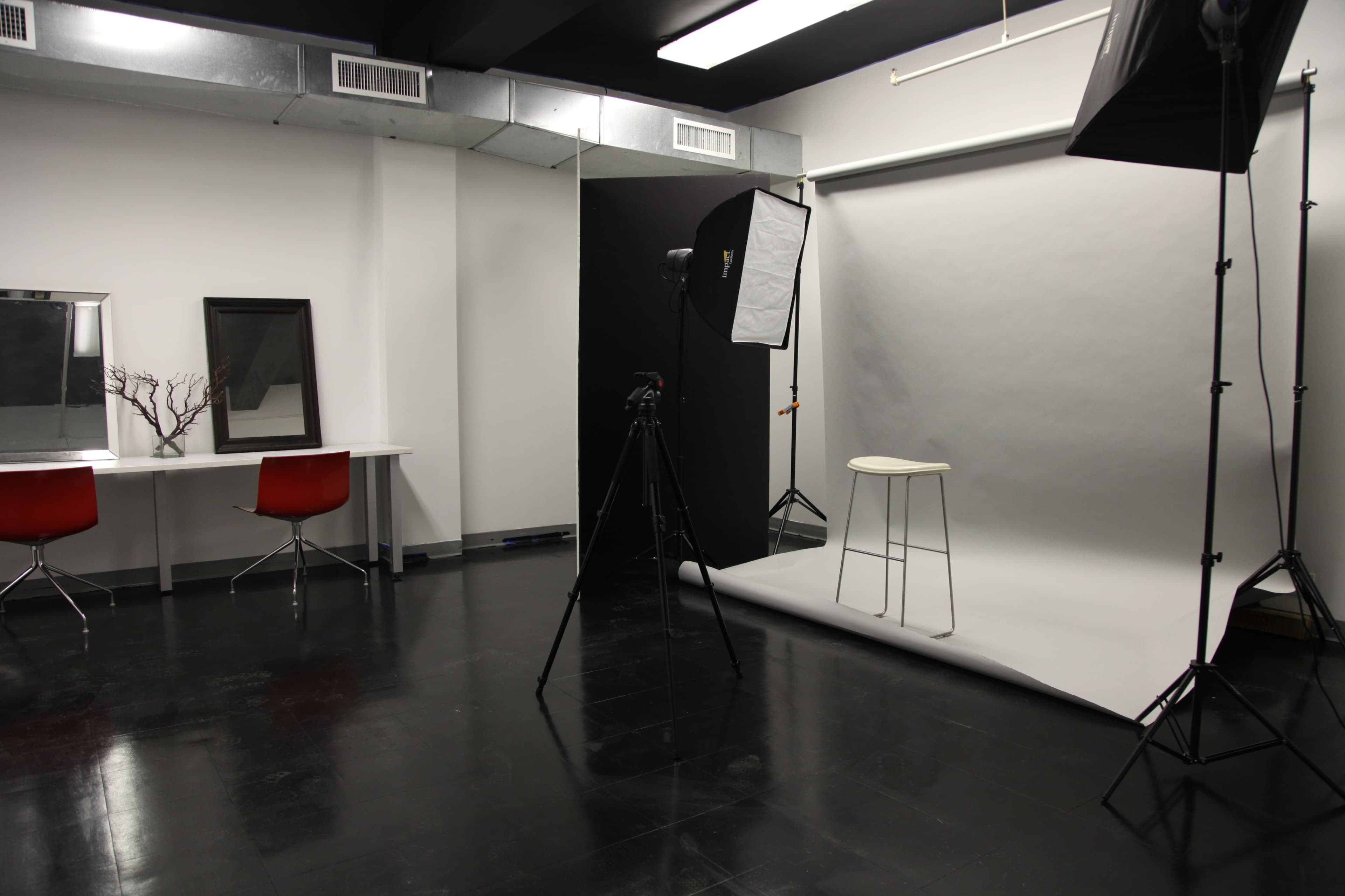 The image shows a photography studio with a gray backdrop, a stool, a camera setup on a tripod, and a mirrored vanity with a red chair.