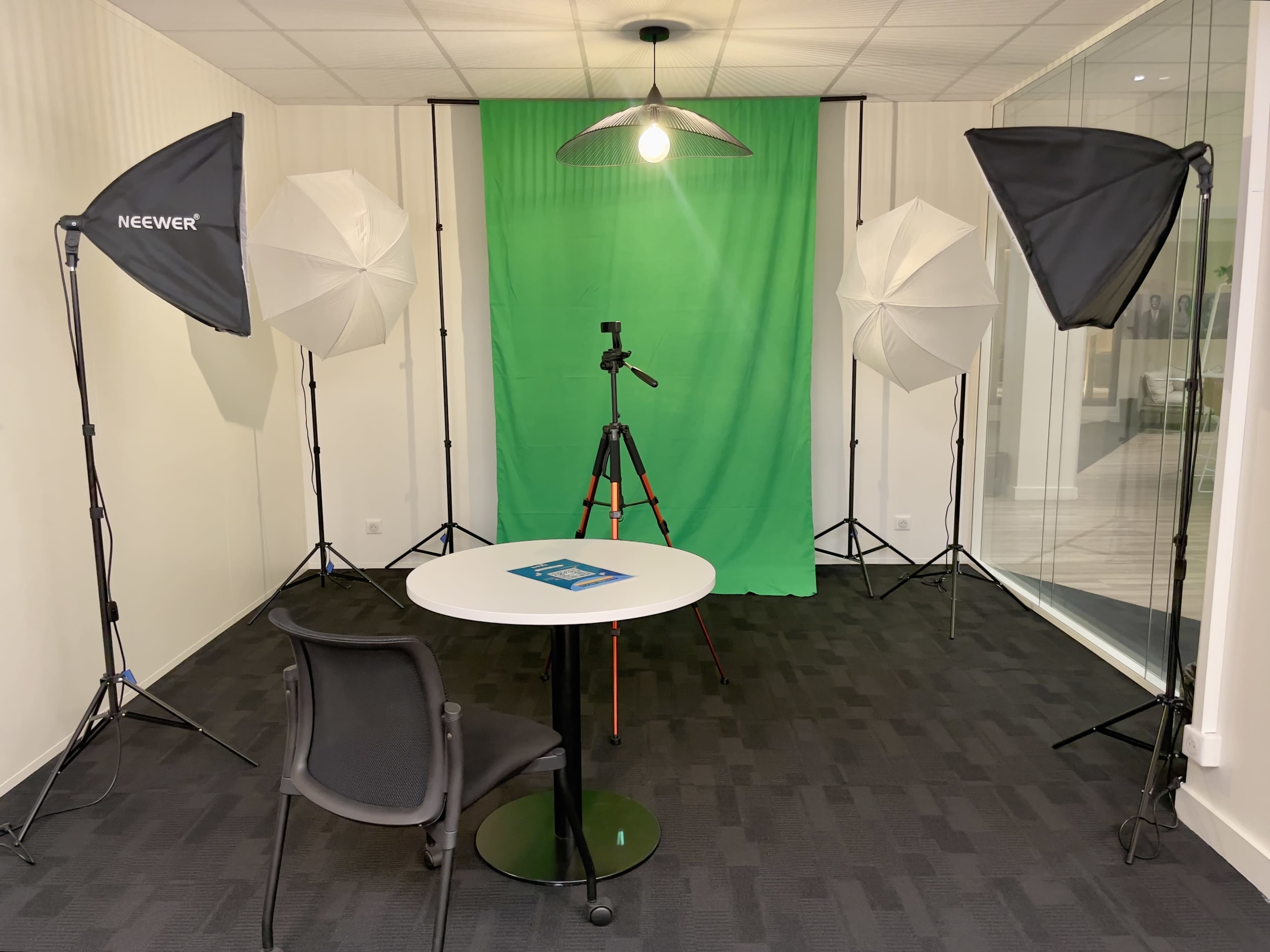 The image shows a small studio setup featuring a green backdrop, a tripod, and two softbox lights surrounding a round table and chair.