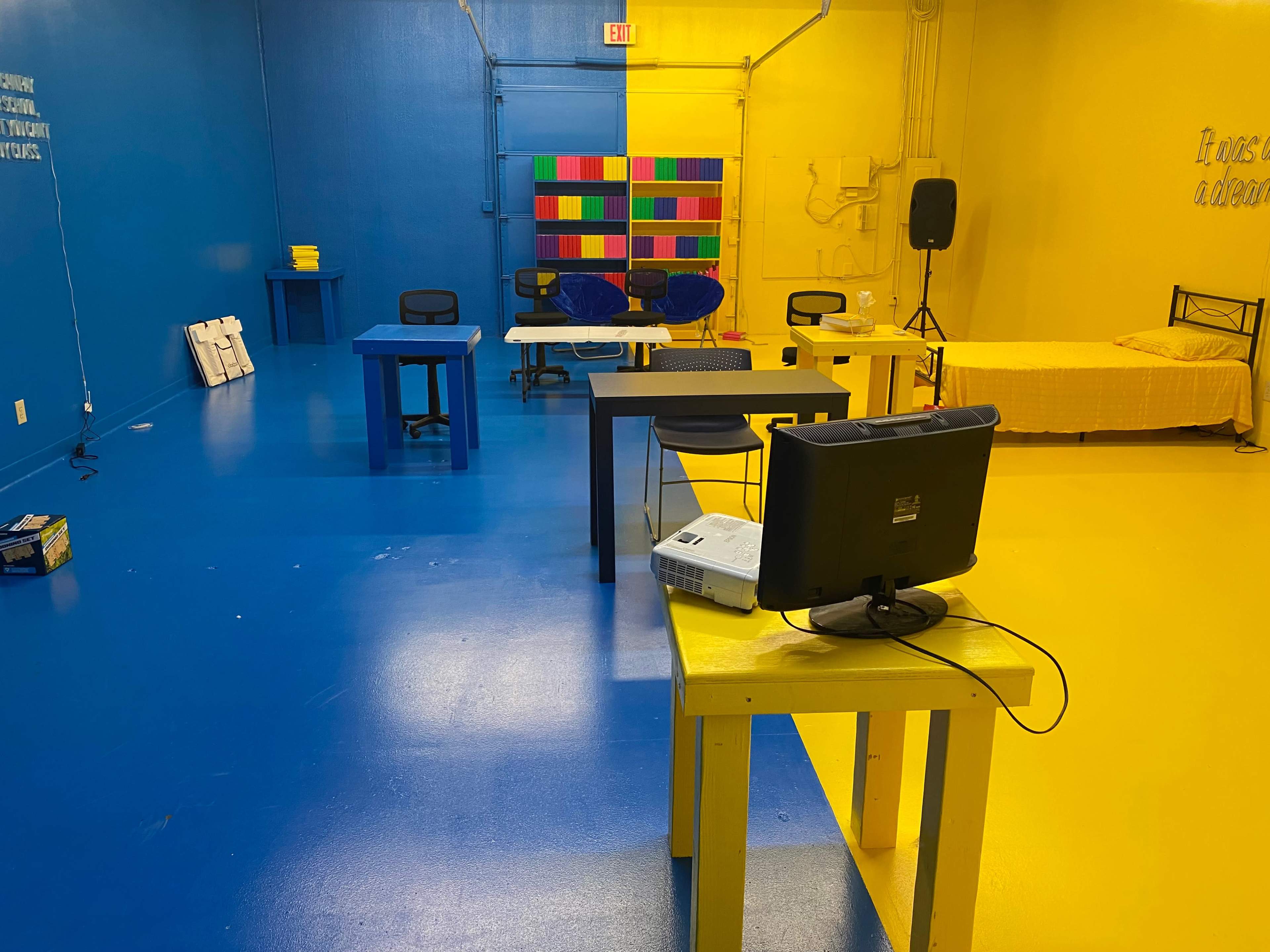 The image shows a brightly colored room with blue and yellow walls, featuring several tables and chairs, a bed, and a projector setup.