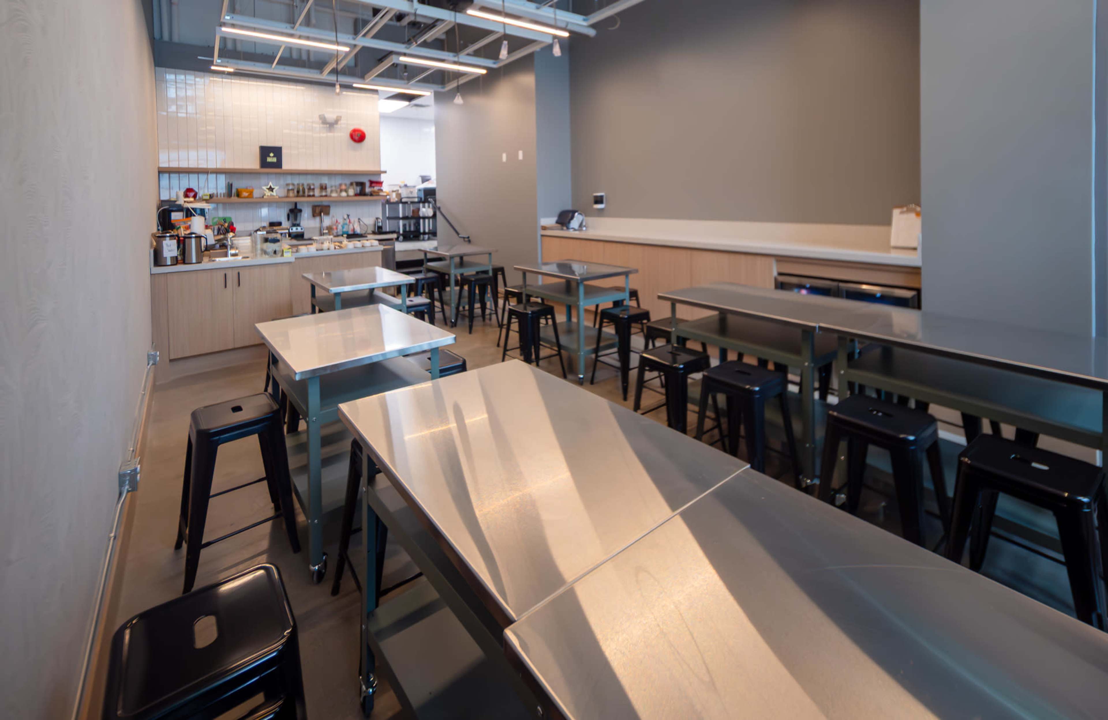 The image shows a modern, industrial-style kitchen space featuring stainless steel tables and stools, with a counter and kitchen equipment in the background.