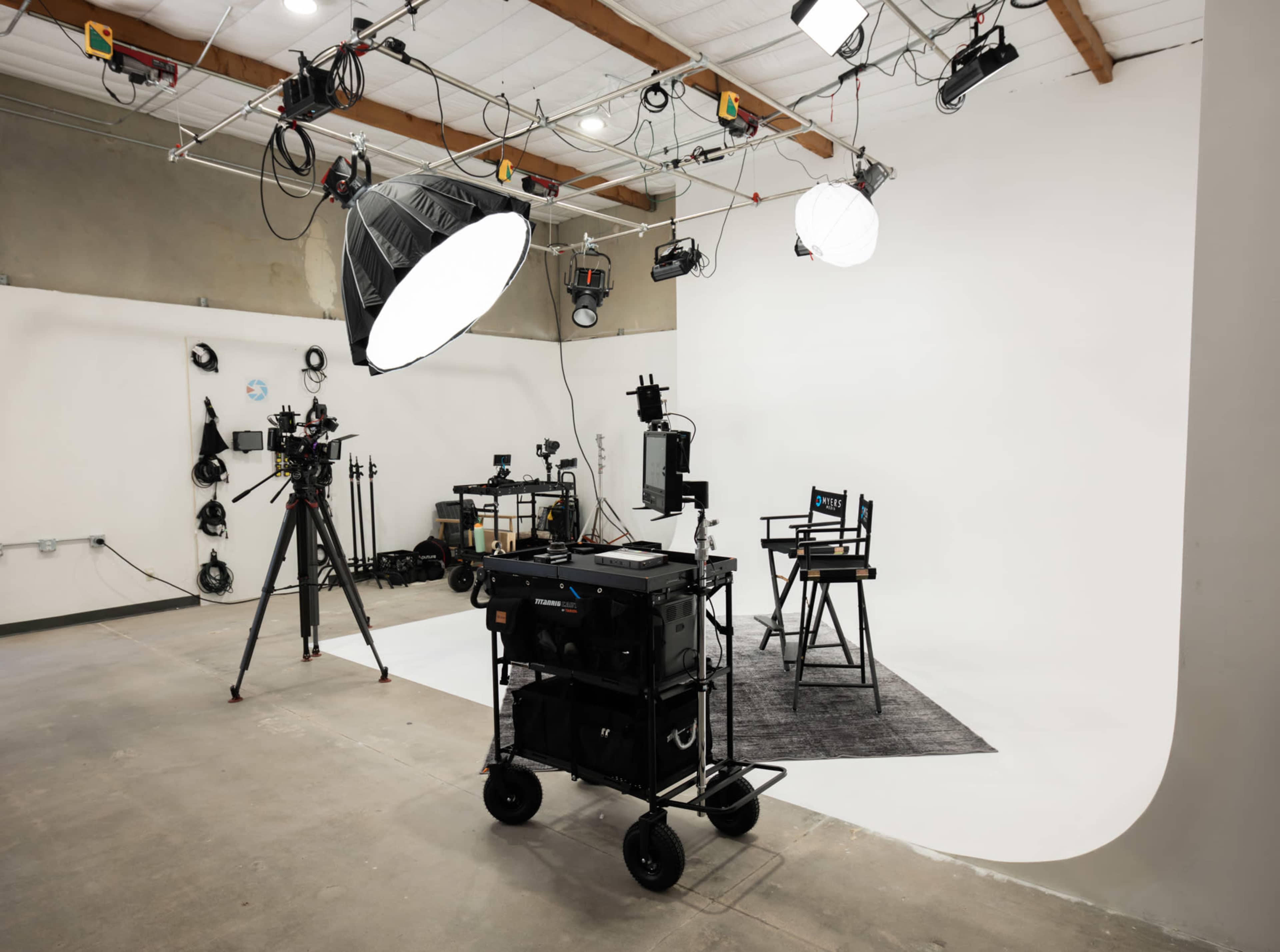 The image shows a photography studio equipped with lighting equipment, a director's chair, and various camera gear arranged on a mobile cart.