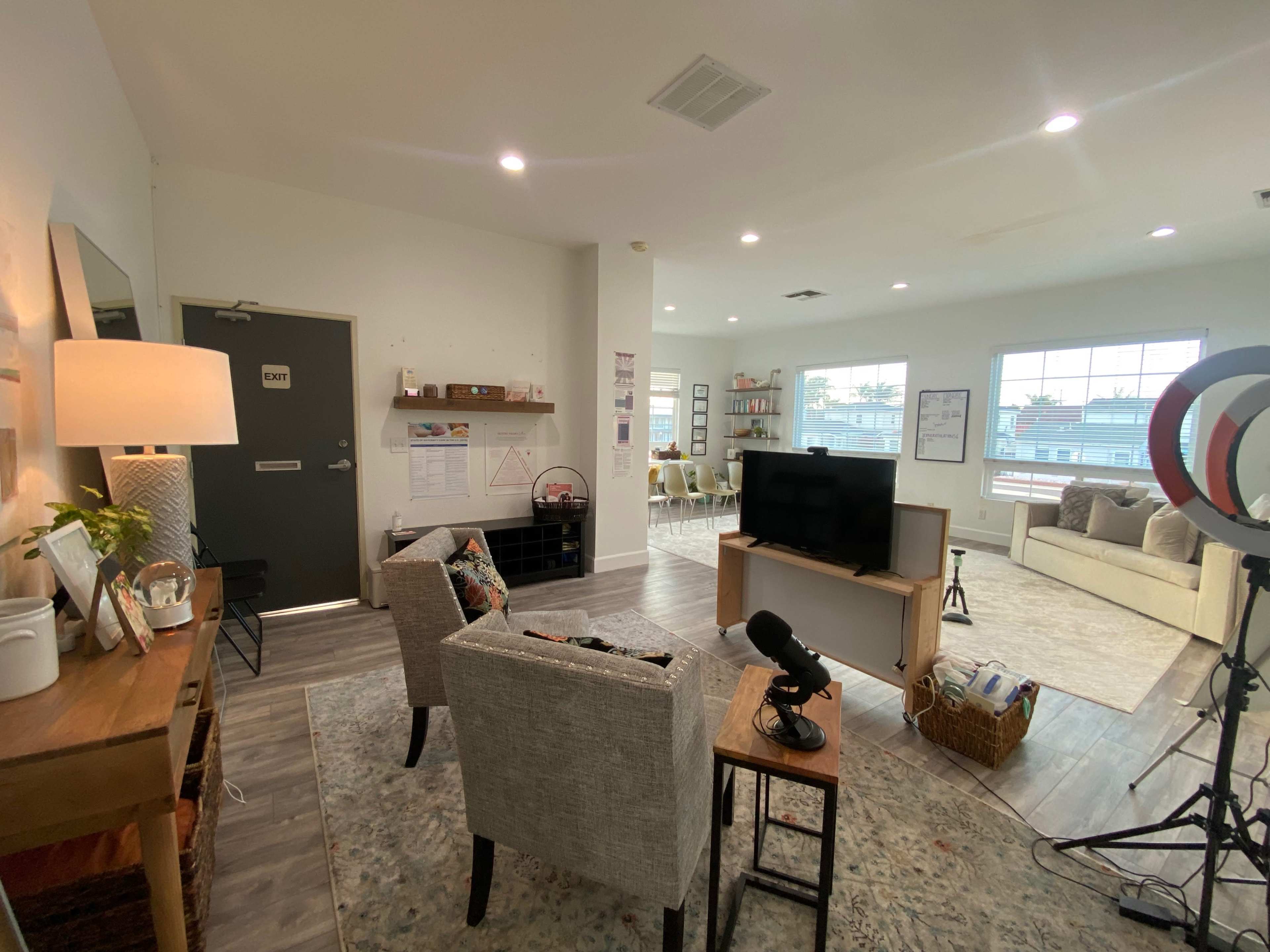 The image shows a cozy living space with a seated area, a television, a desk with a lamp, and large windows allowing natural light.