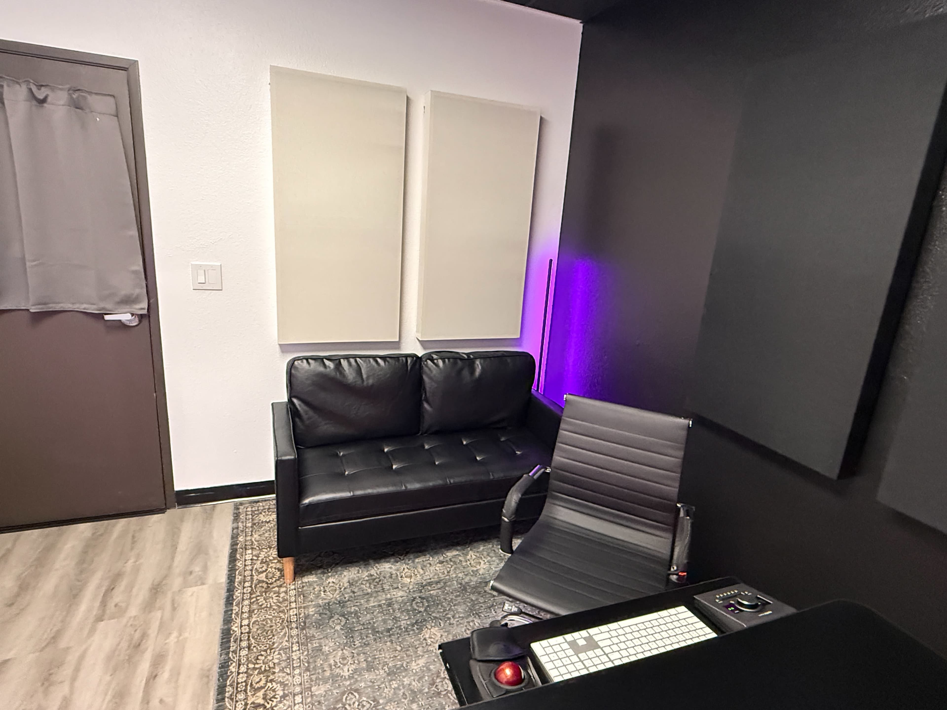 The image shows a small, minimalist room with a black couch, a modern chair, a desk with equipment, and soundproofing panels on the walls.