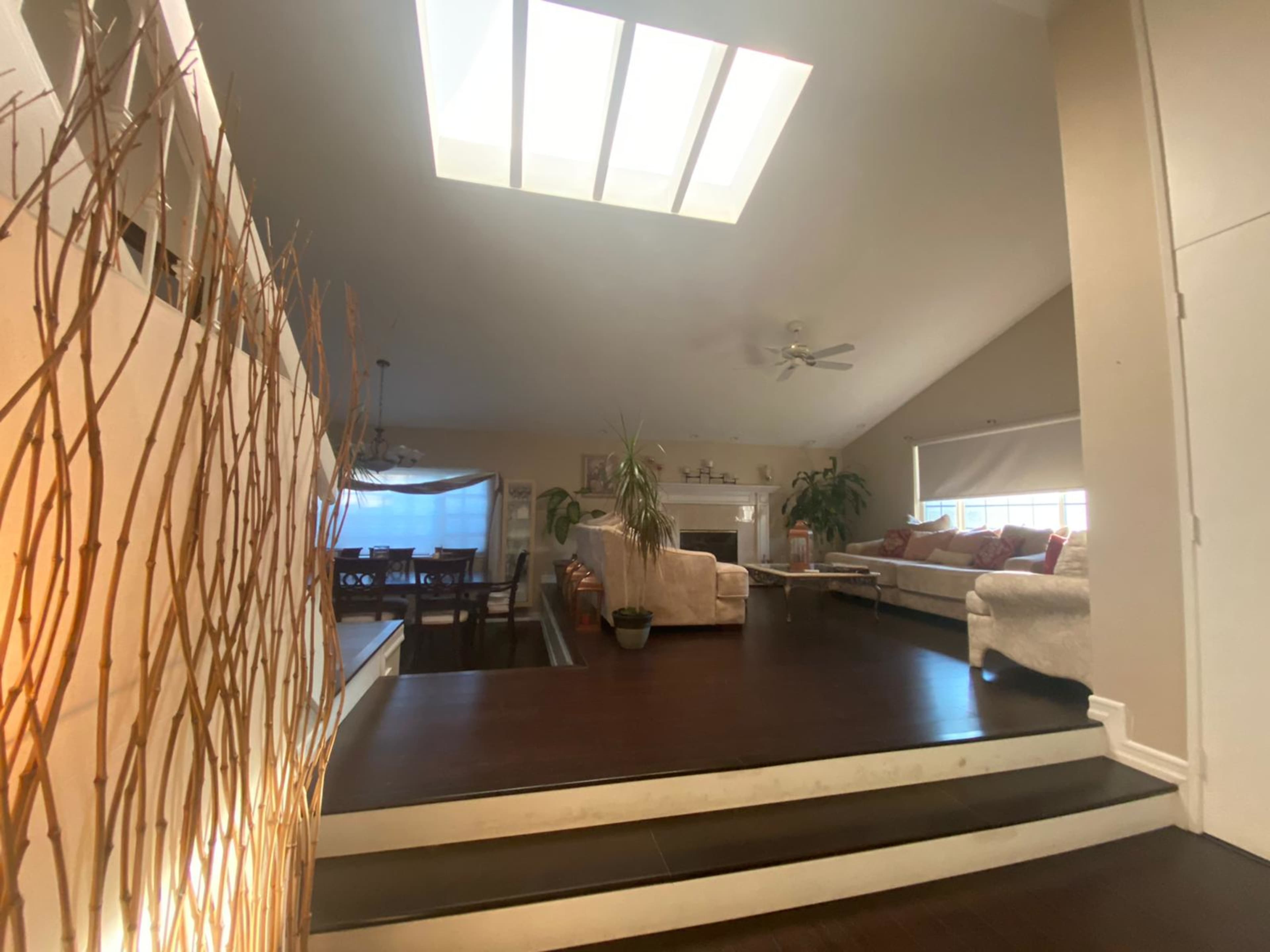 A spacious living area with a skylight, hardwood floors, and a staircase leading to an elevated section featuring a couch and plants.