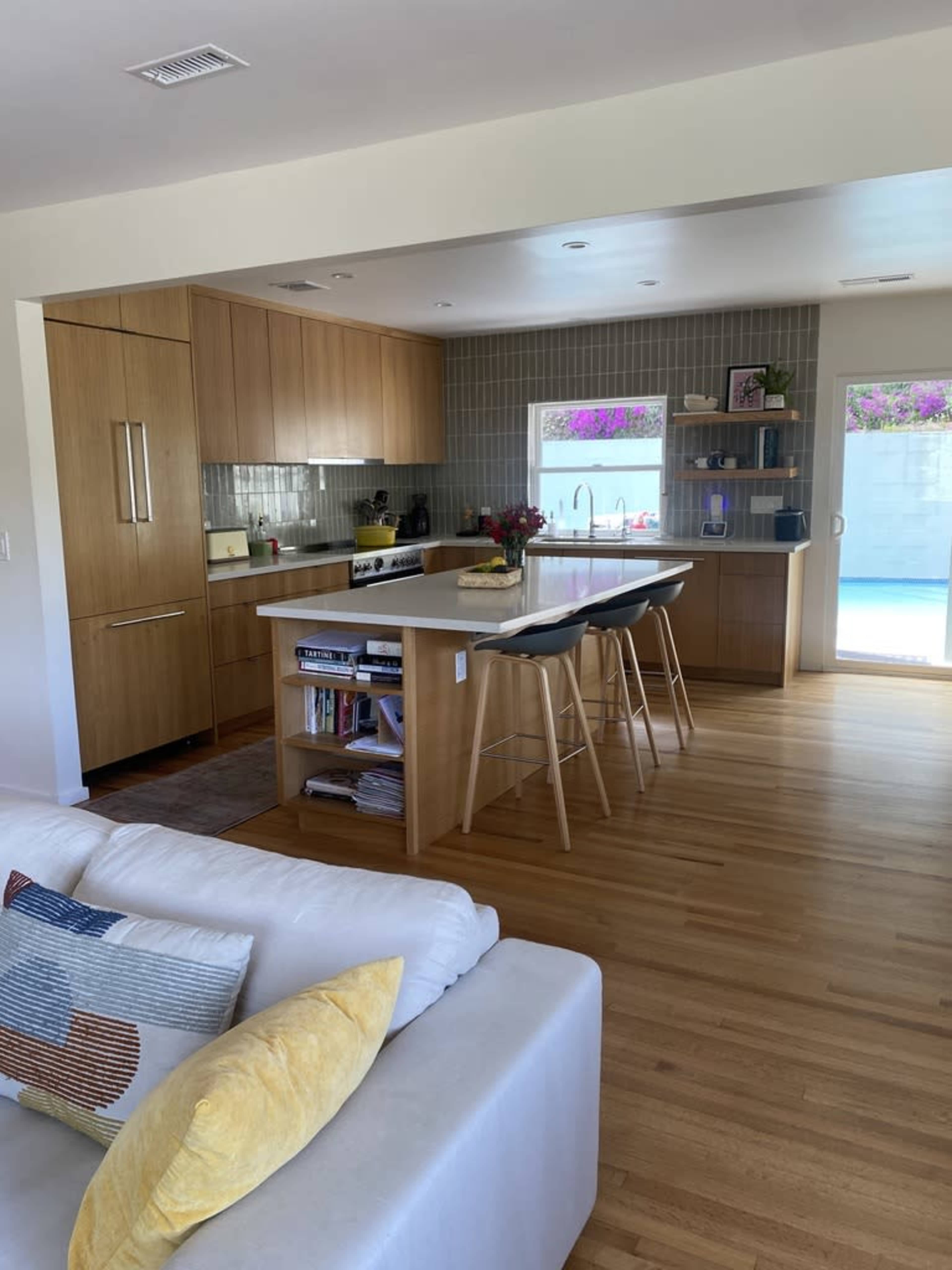 The image shows a modern kitchen with wooden cabinetry, a central island with bar stools, and a view of a pool through a sliding glass door.