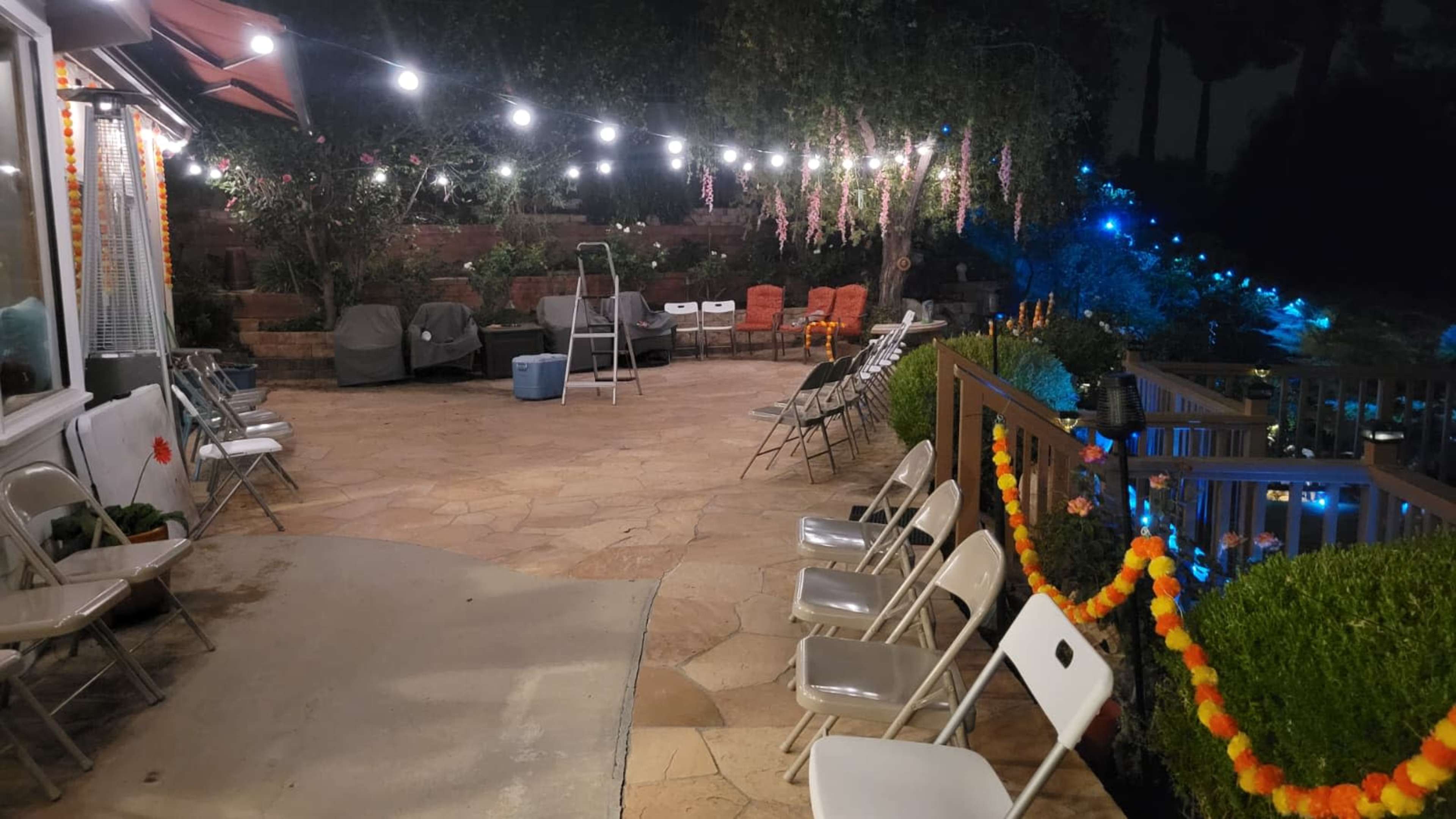 The image shows a well-lit outdoor patio with folding chairs arranged in rows, surrounded by greenery and decorative lights.