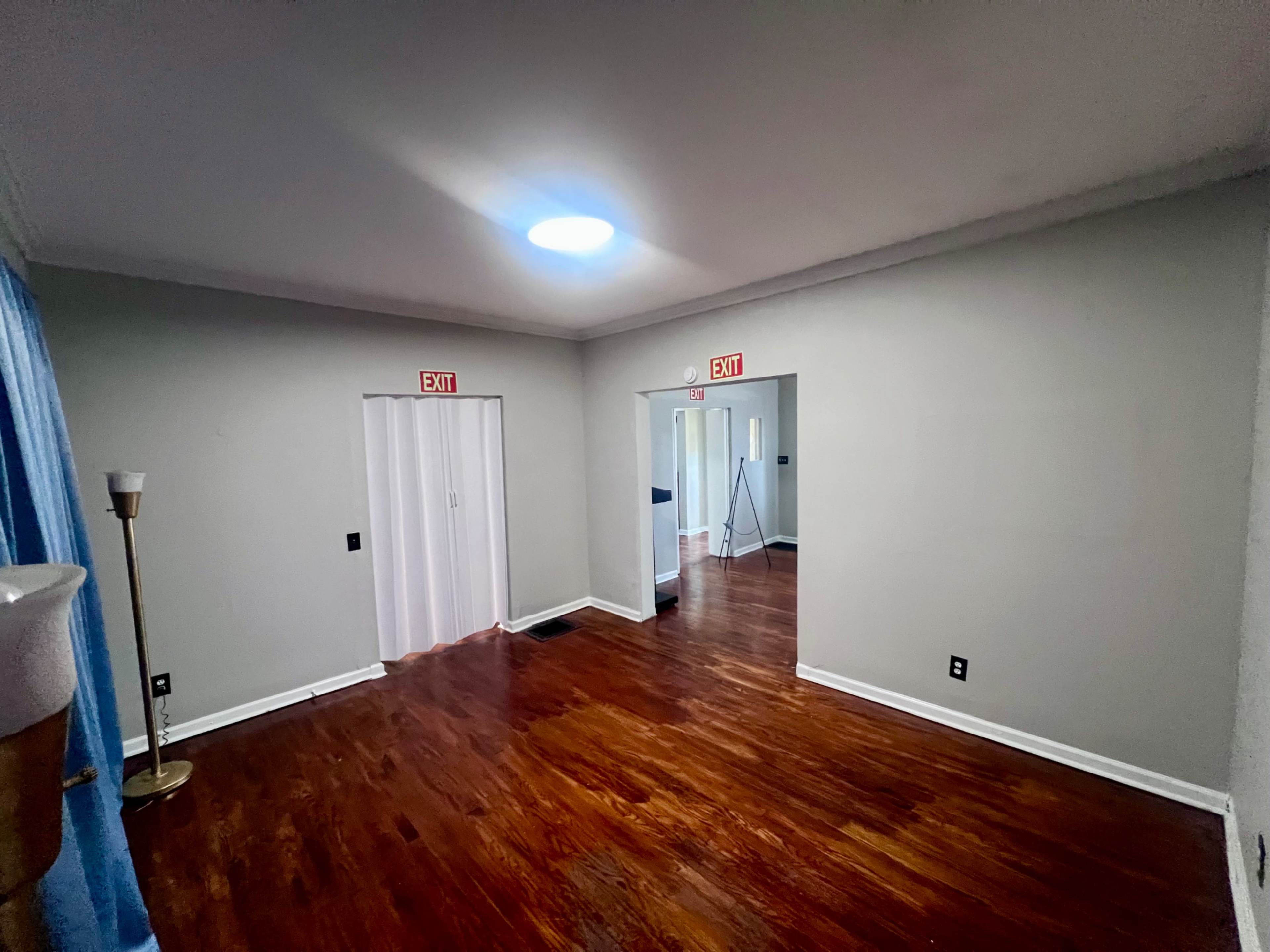 The image shows a sparsely furnished room with wooden floors, a ceiling light, and two exits marked with "EXIT" signs.