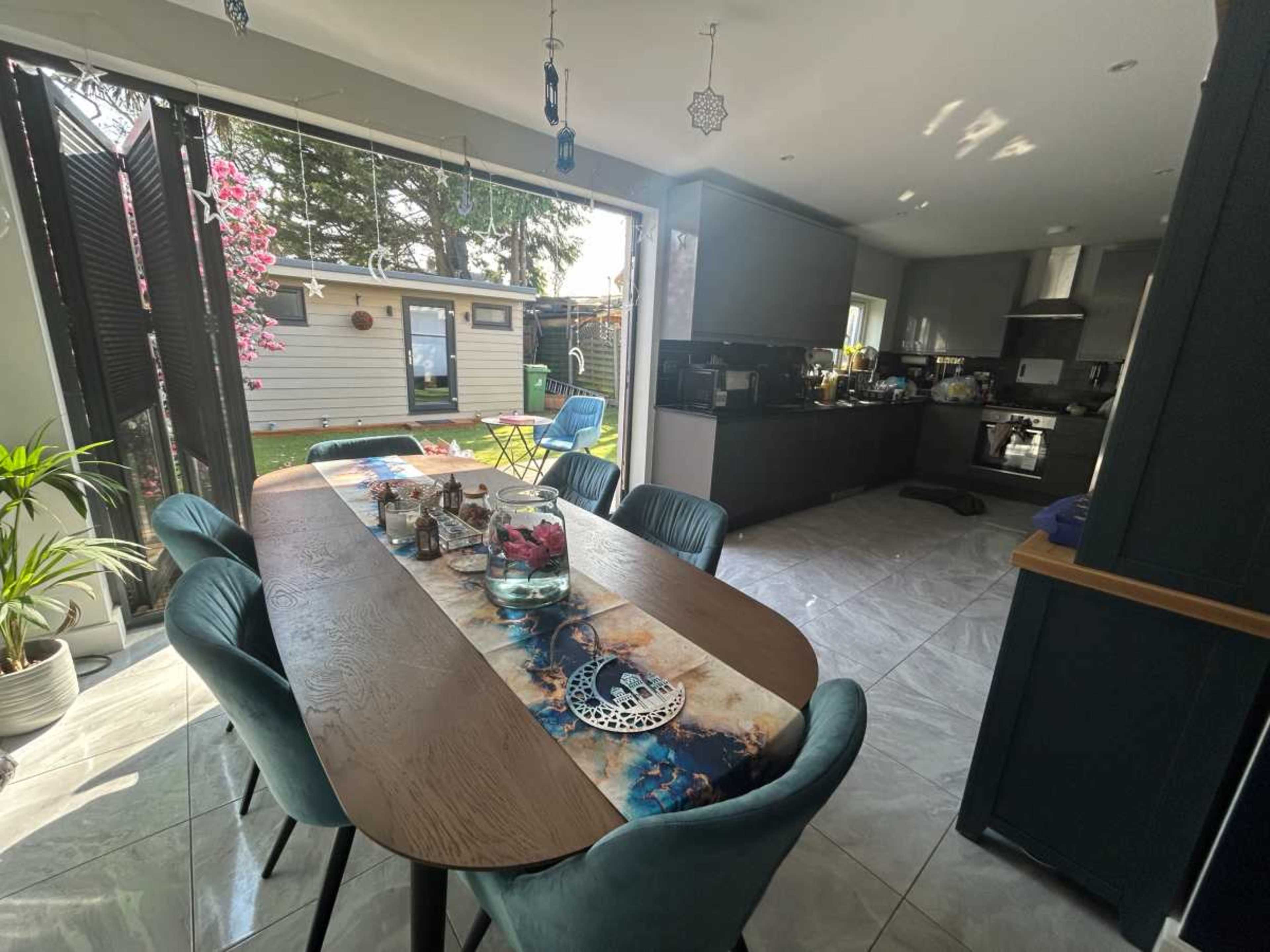 A modern dining area features a large wooden table with blue chairs, surrounded by large windows that open to a garden.