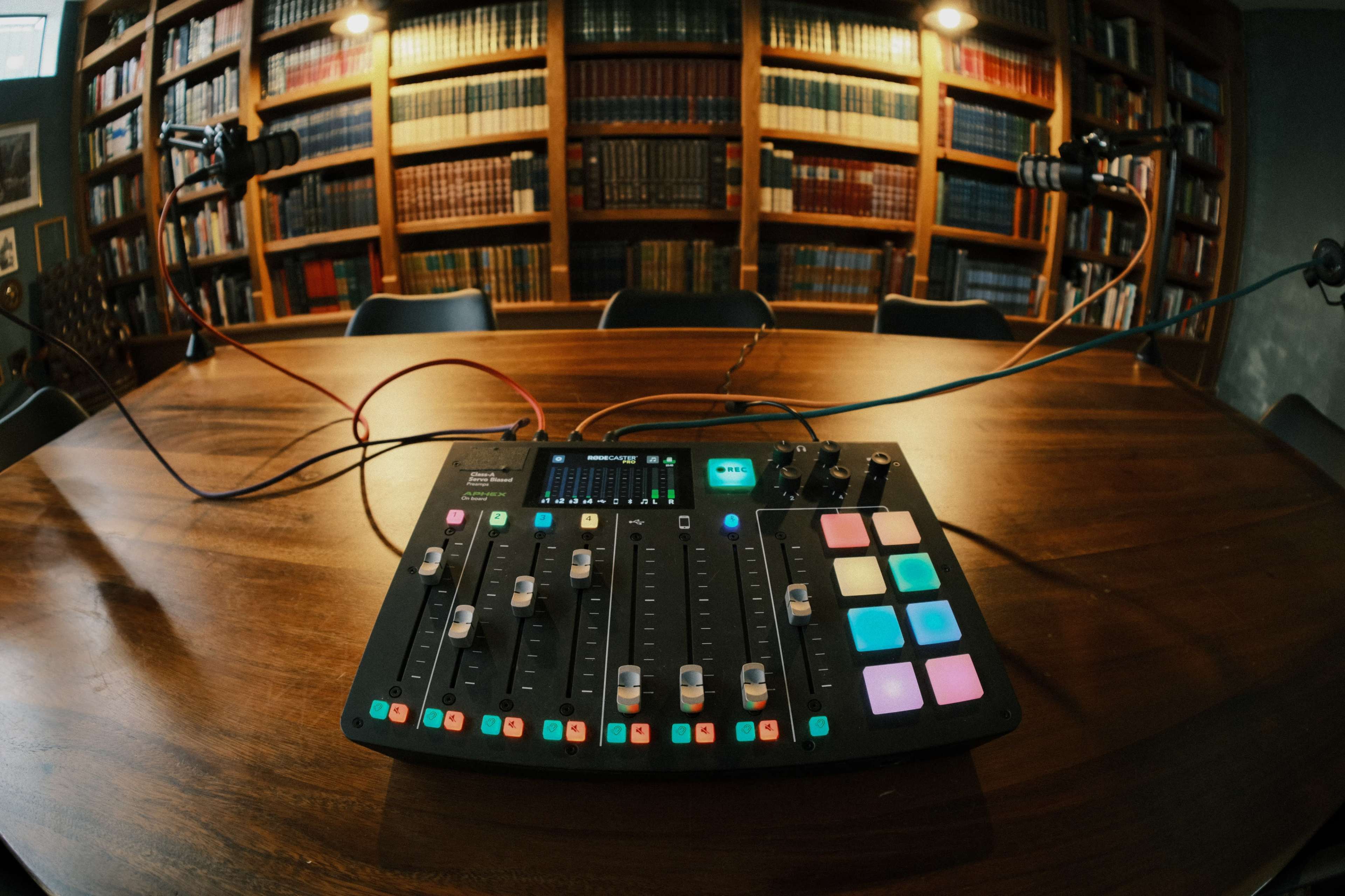 A mixing console is placed on a wooden table in a room lined with bookshelves.