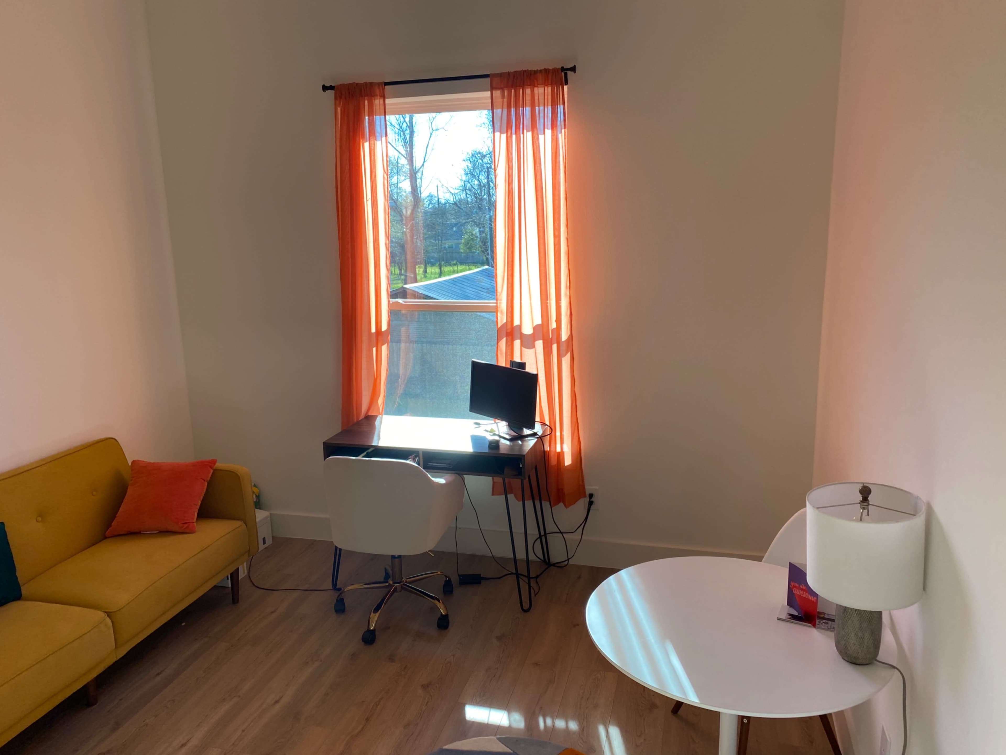 A bright room with a yellow sofa, a white table, a desk with a computer, and large orange curtains over a window.