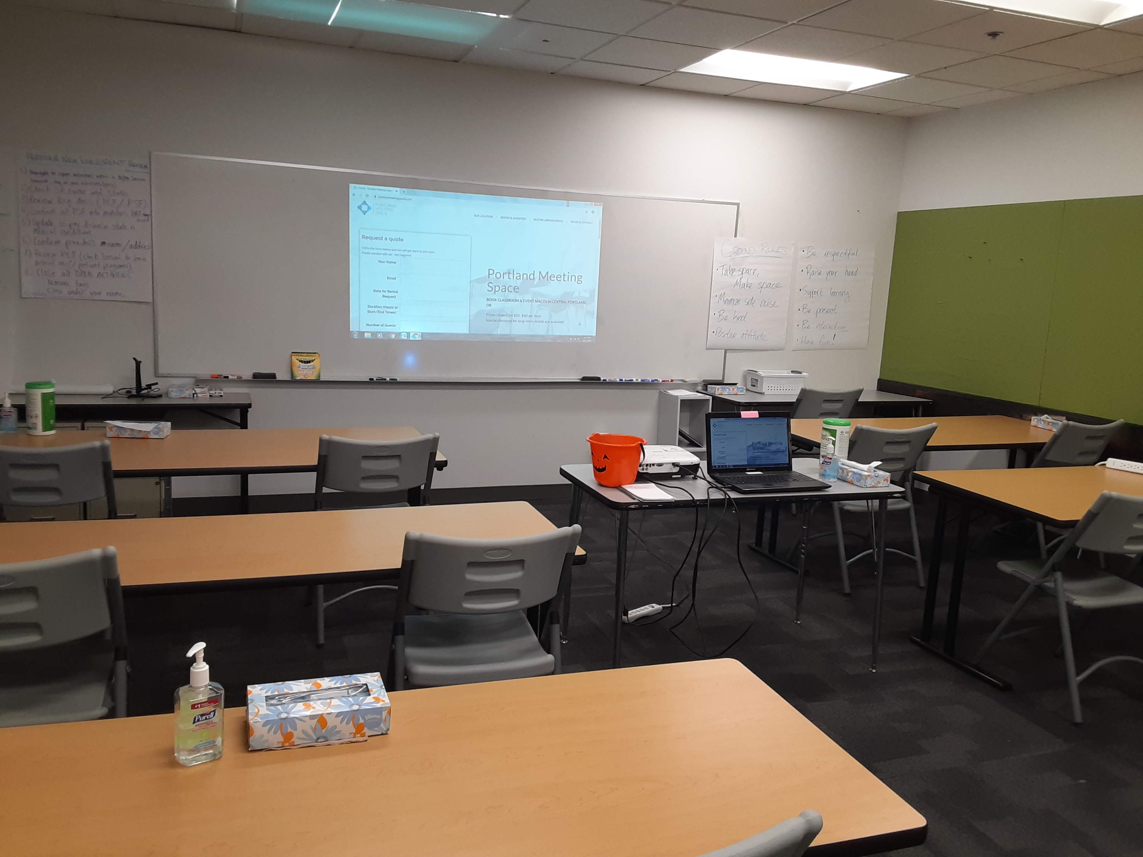 A classroom is set up with tables and chairs arranged for a meeting, featuring a projector screen and various supplies on the tables.