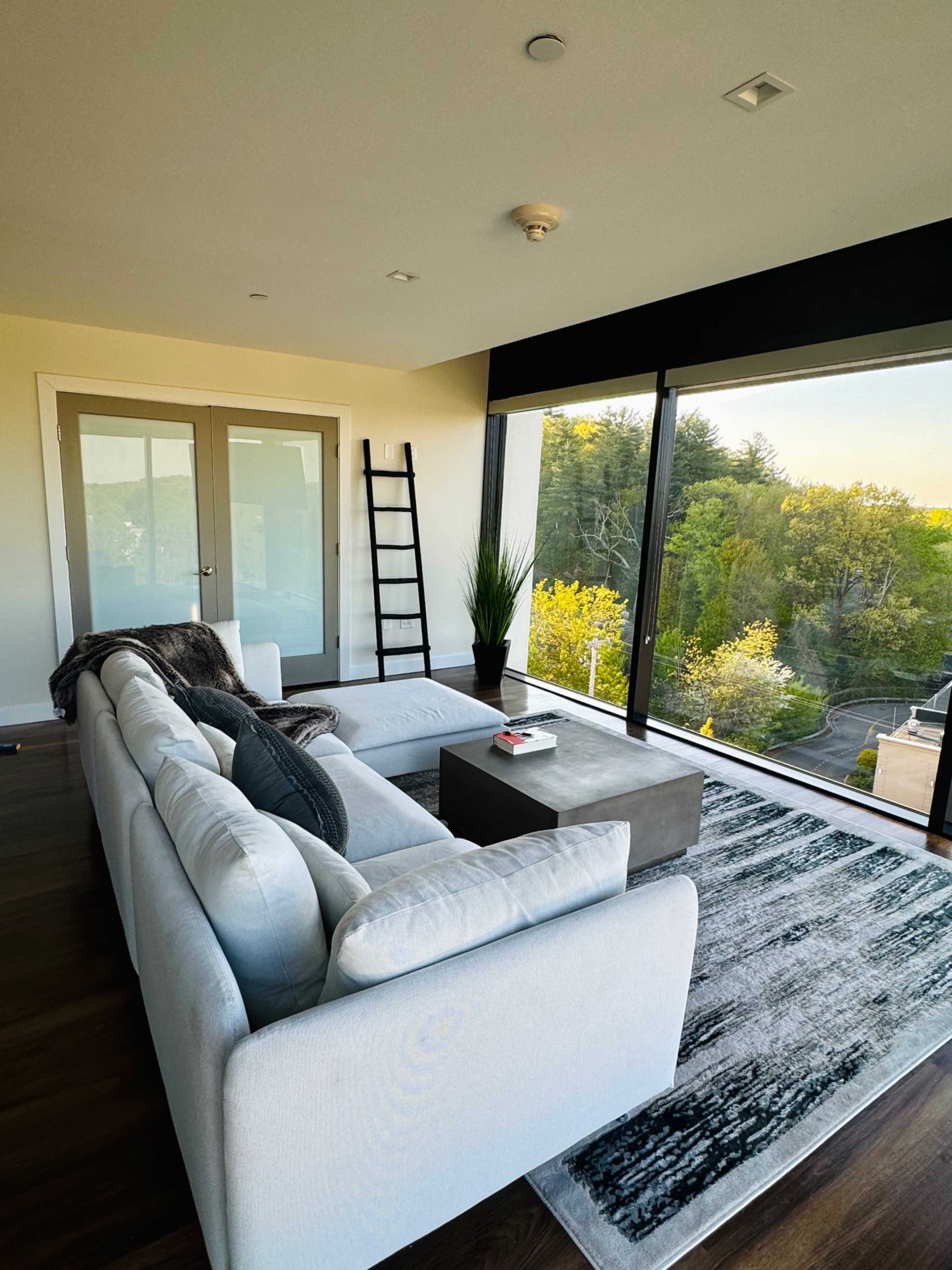A modern living room features a large sectional sofa, a small coffee table, and floor-to-ceiling windows offering a view of greenery outside.