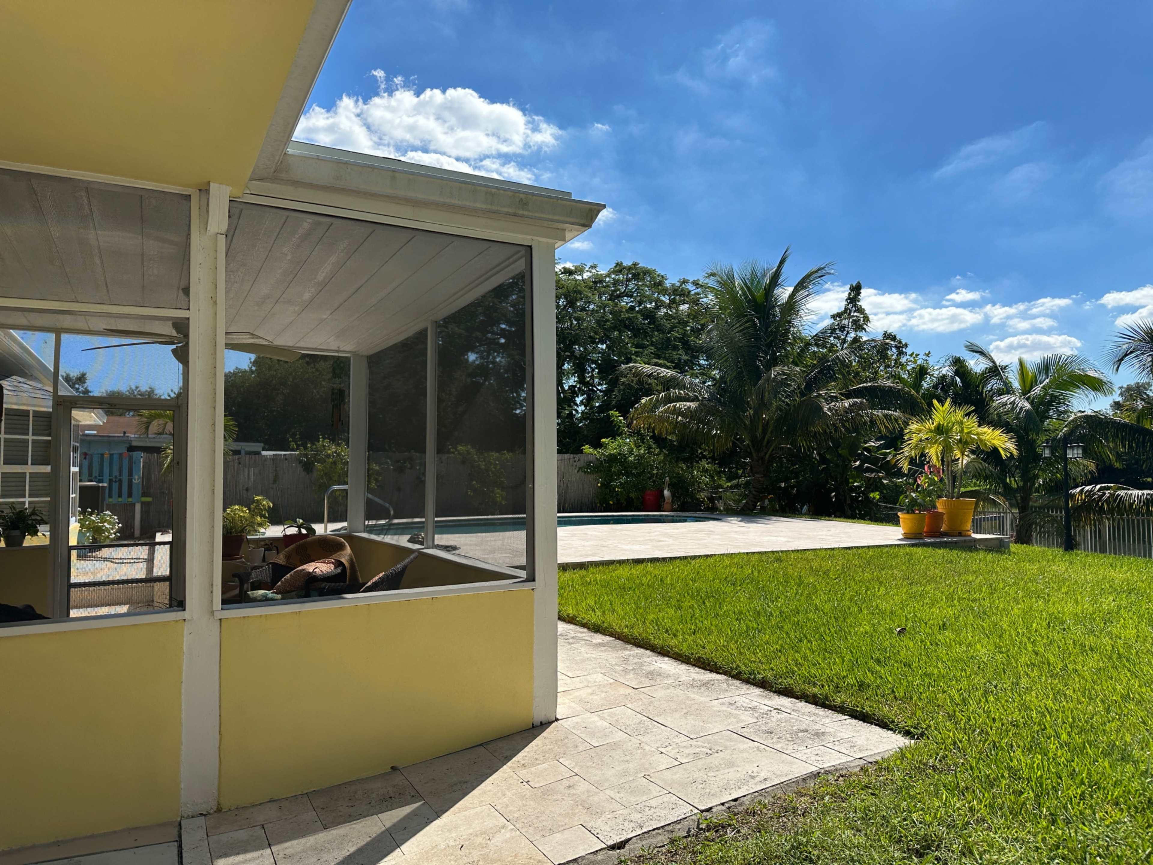 A house with a screened porch overlooks a backyard featuring a pool and landscaped greenery under a bright blue sky.