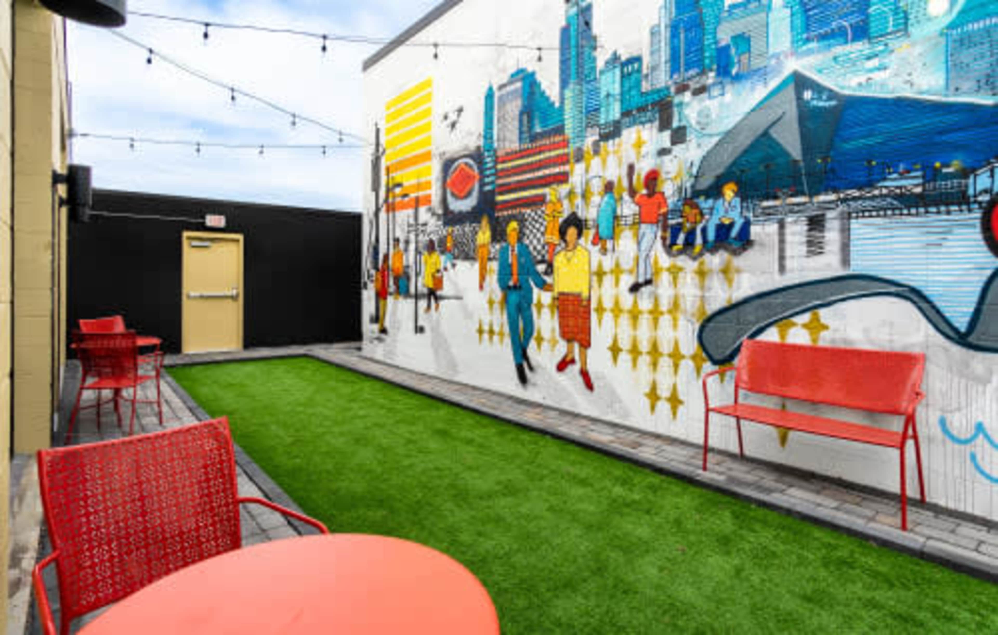 A colorful mural on a wall in an outdoor space with artificial grass, red chairs, and tables.