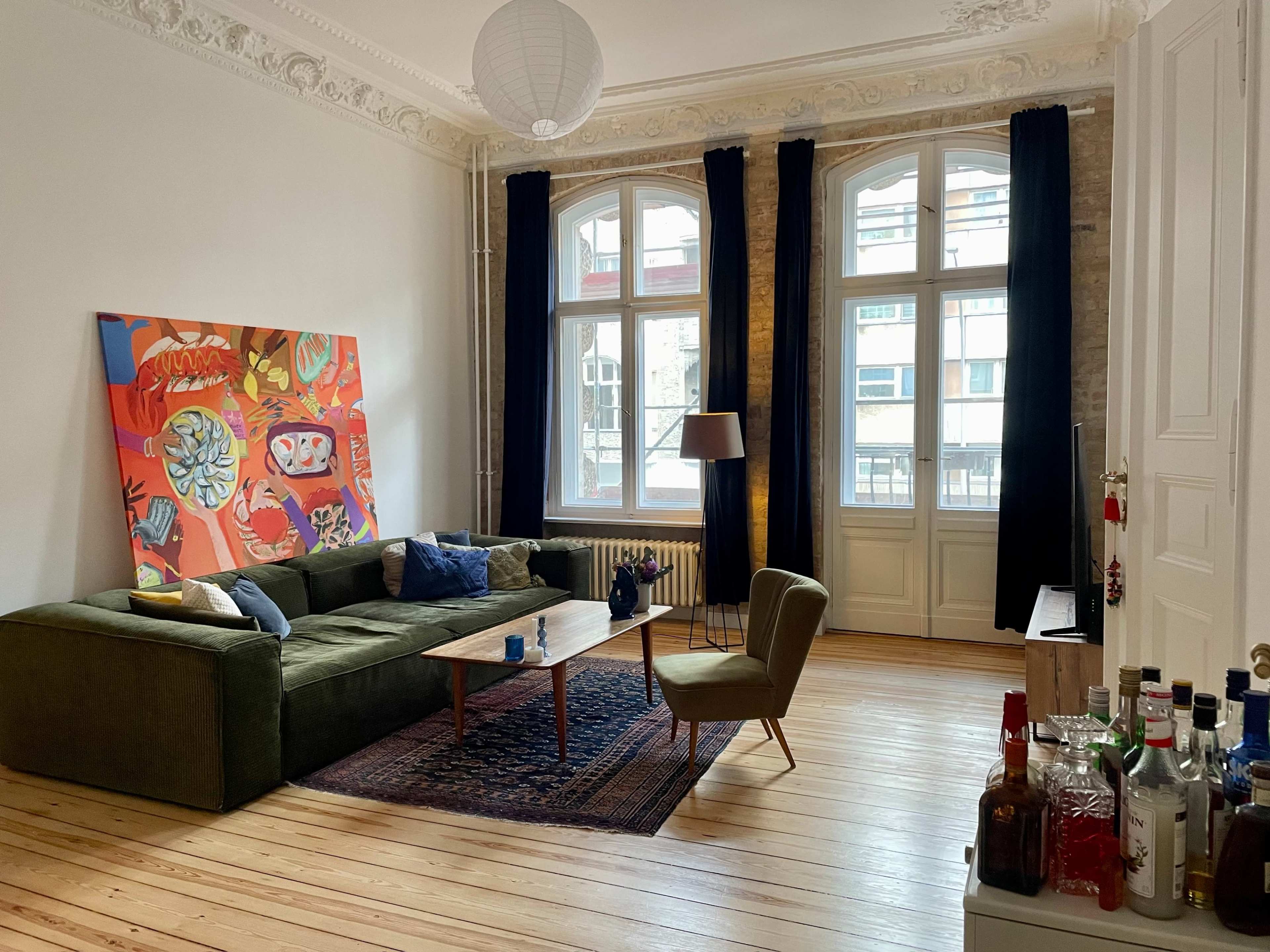 A bright living room with large windows, a green sectional sofa, a wooden coffee table, and an abstract painting on the wall.