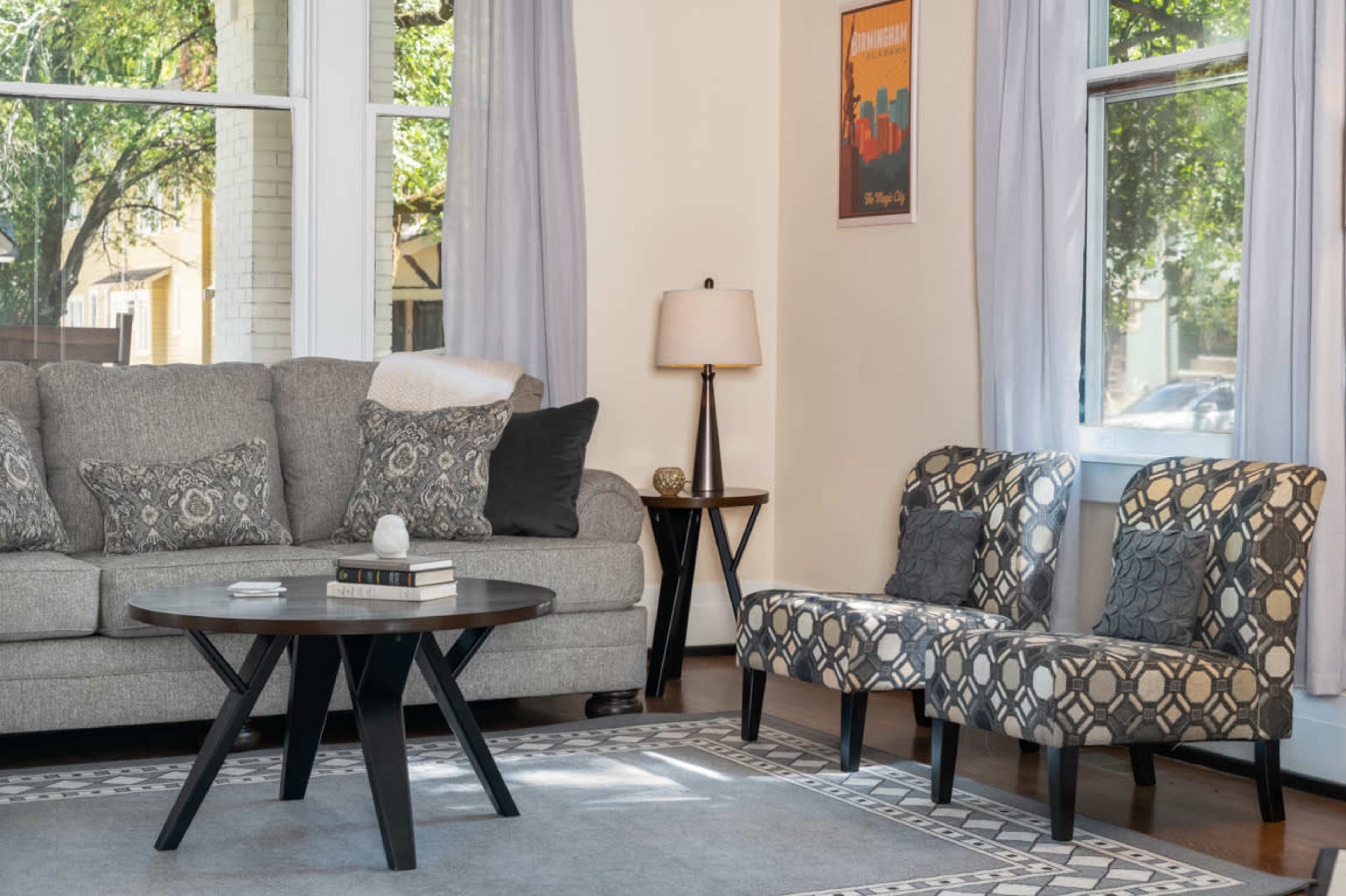 A cozy living room features a gray sofa, two patterned chairs, a circular coffee table, and a lamp beside a window.