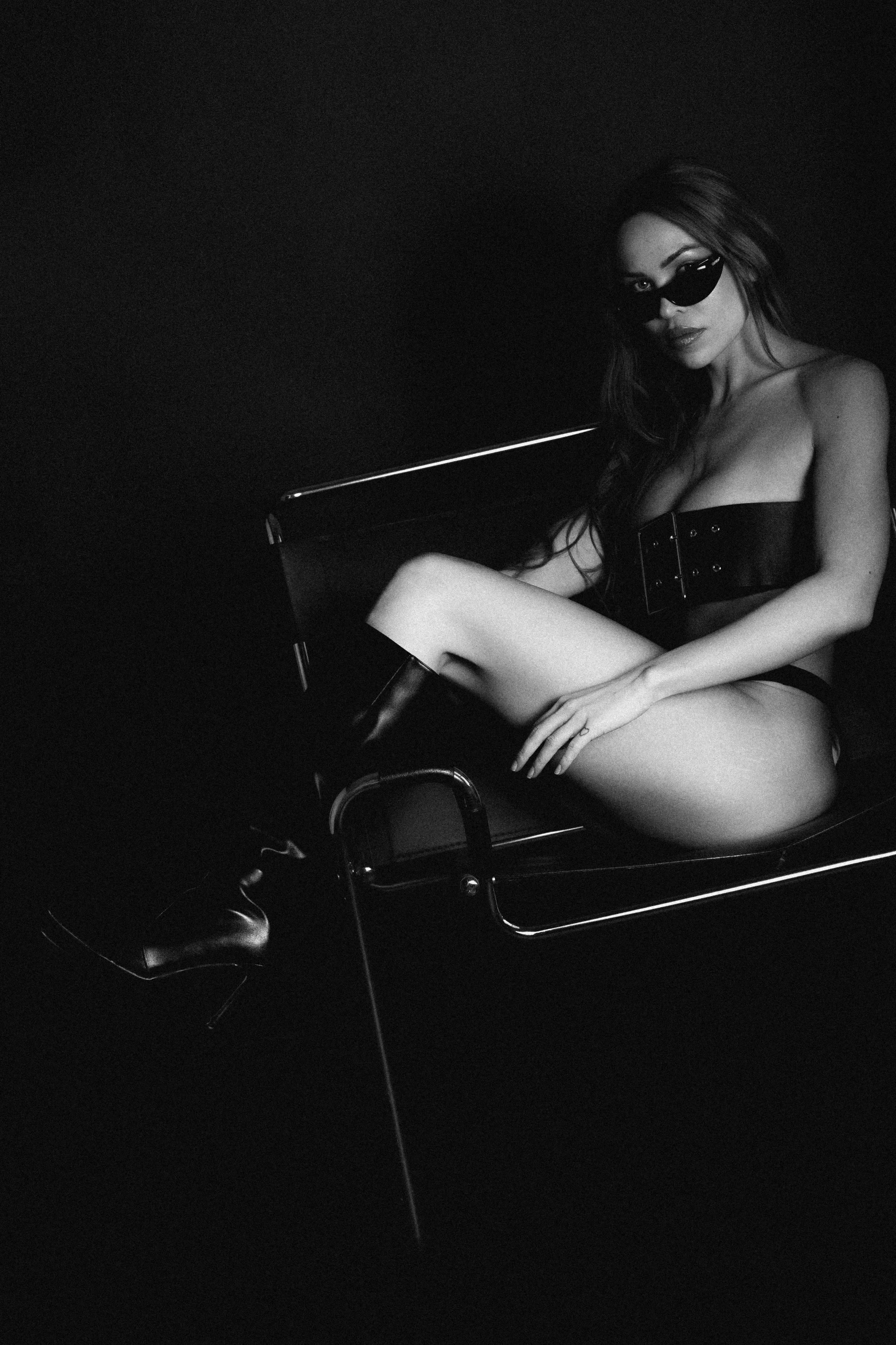 A person wearing sunglasses and a belt sits on a chair in a dimly lit space.