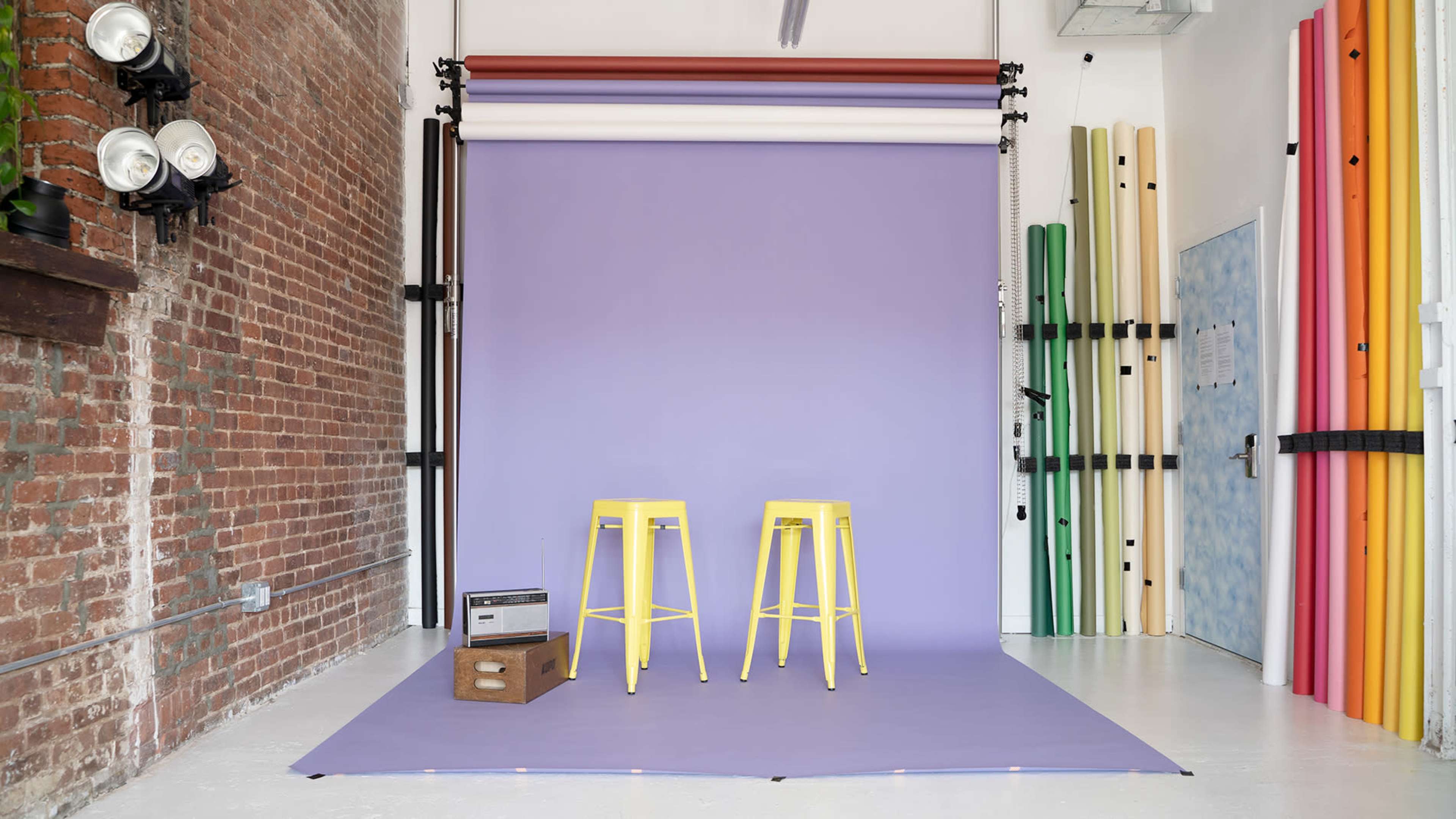 A photography studio features two yellow stools in front of a purple backdrop, with various rolled backdrops visible on the sides.