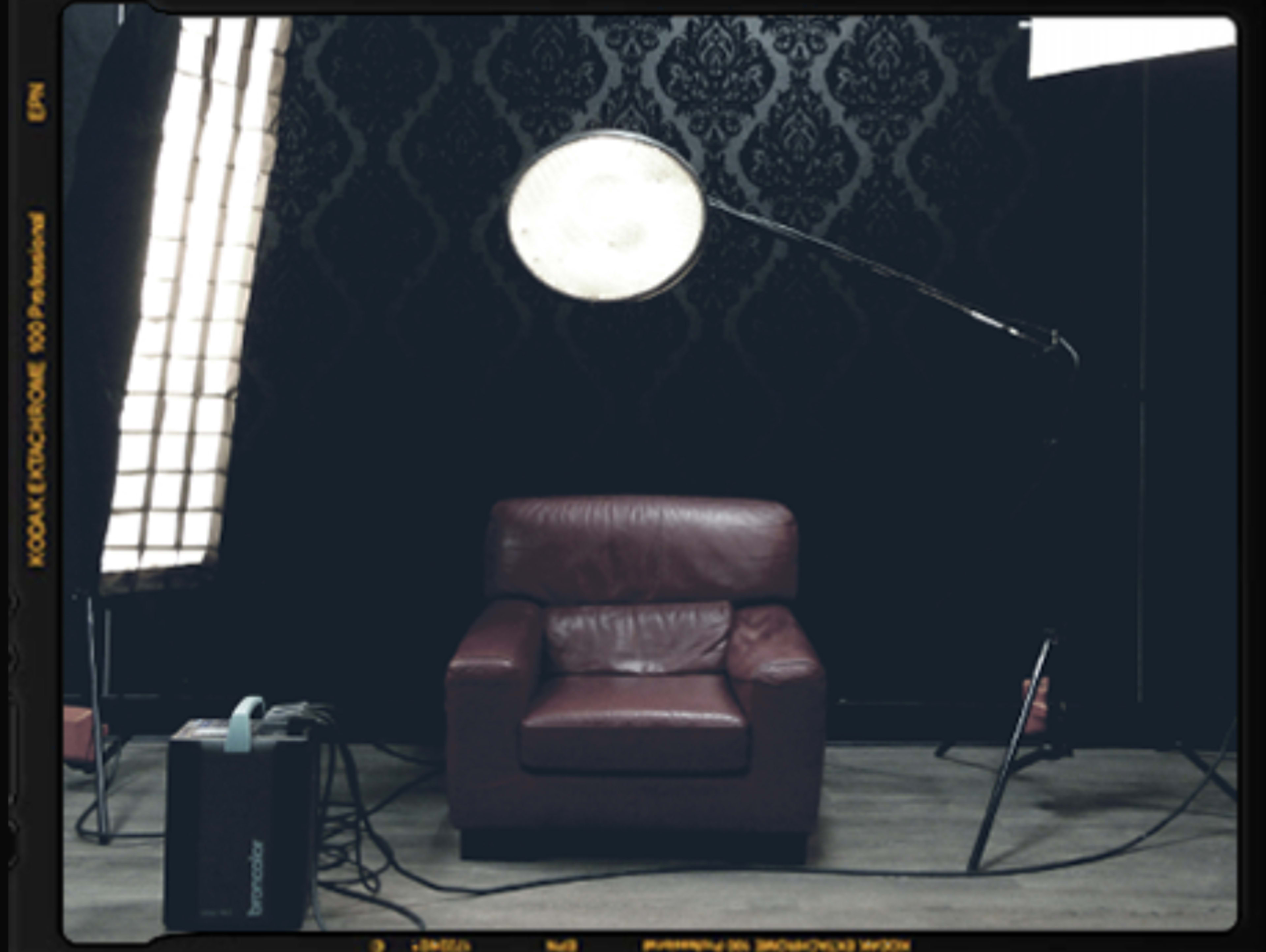 A burgundy armchair is positioned in front of a textured black wall, with studio lighting equipment arranged nearby.