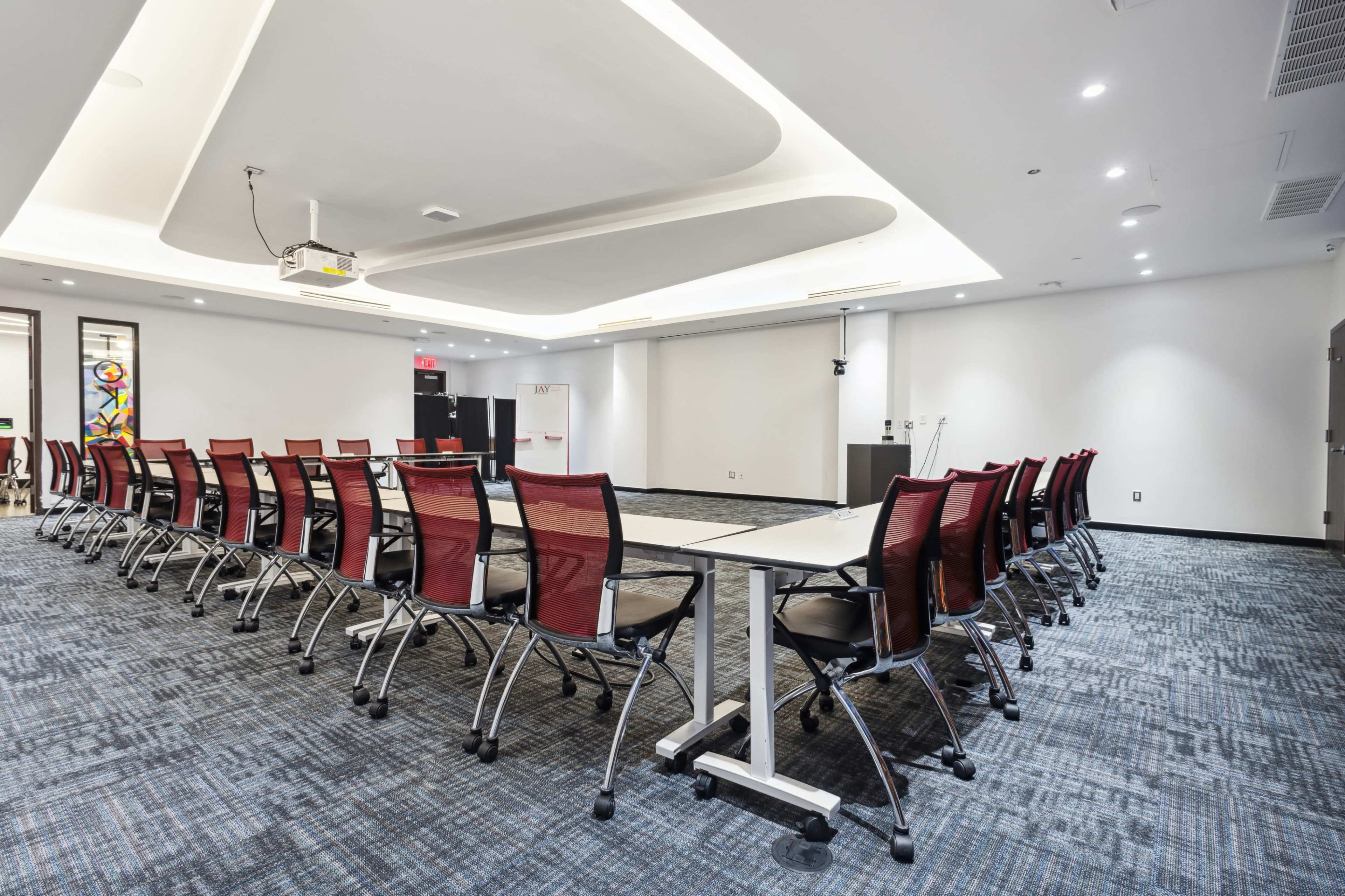 A boardroom is arranged with a long table surrounded by red mesh chairs on a gray carpet.