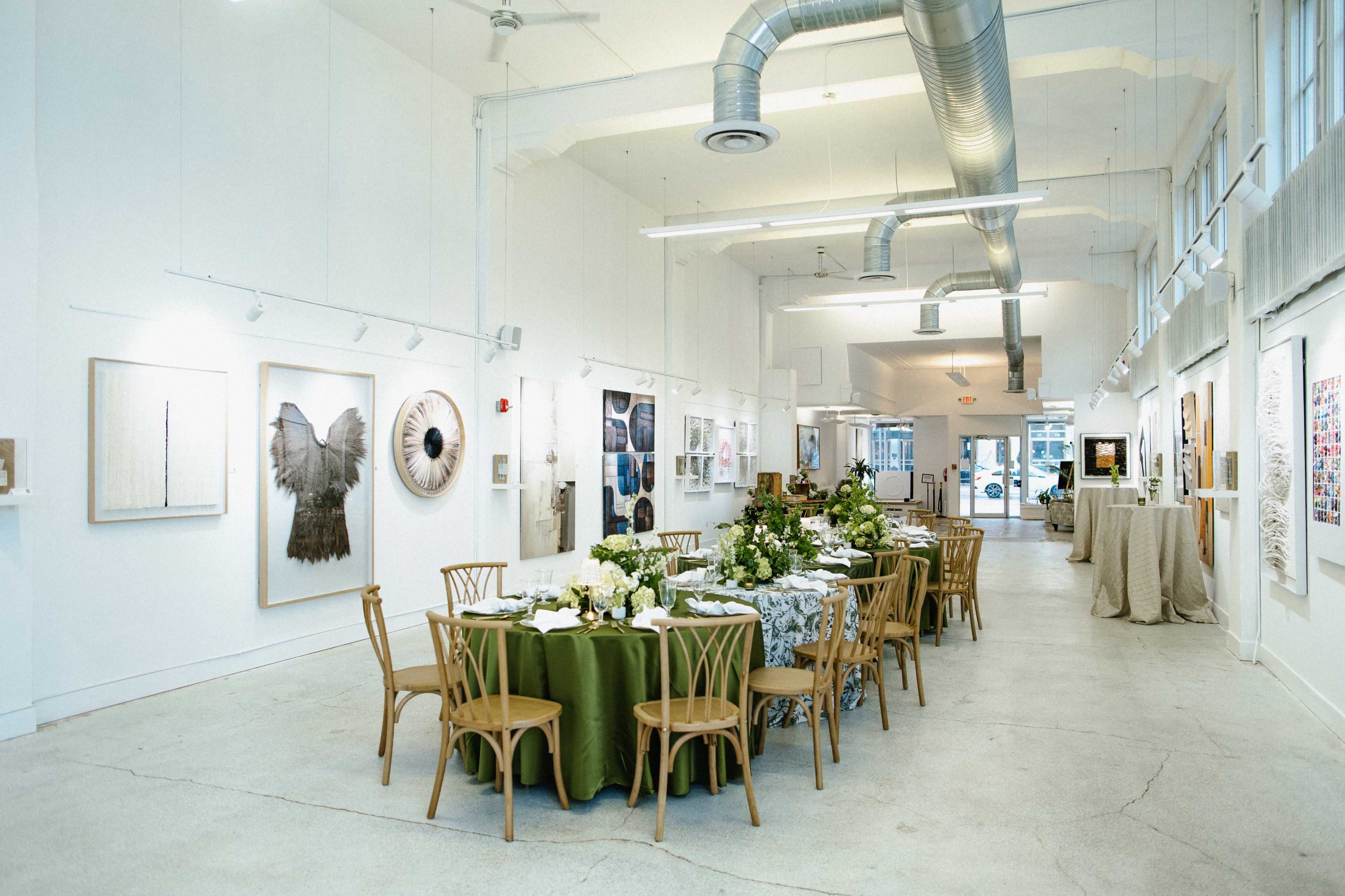 The image shows a spacious gallery with a long dining table set for an event, surrounded by various pieces of artwork displayed on the white walls.