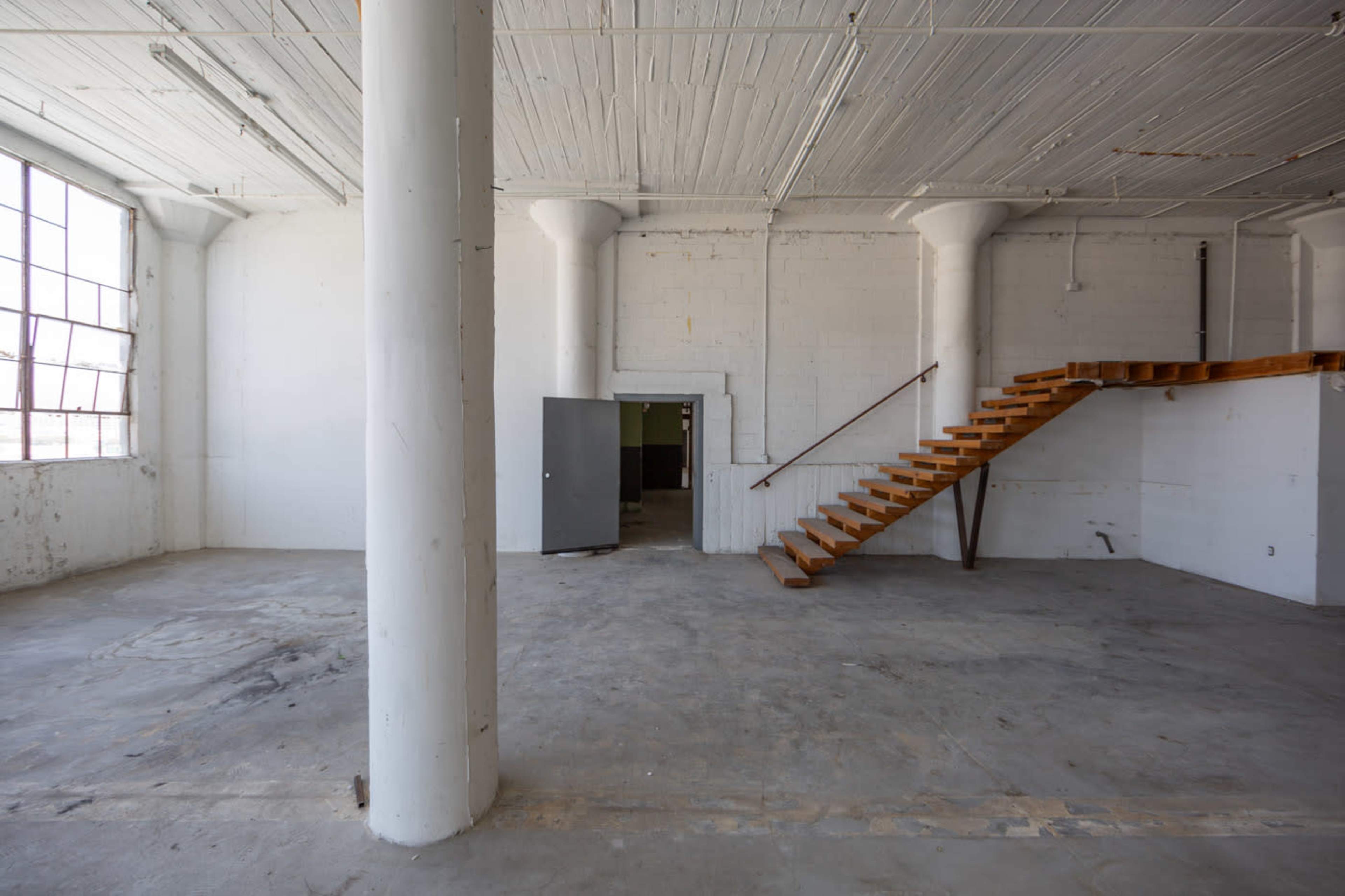 A spacious, unfinished industrial room with concrete floors, large windows, and a wooden staircase leading to a small upper area.