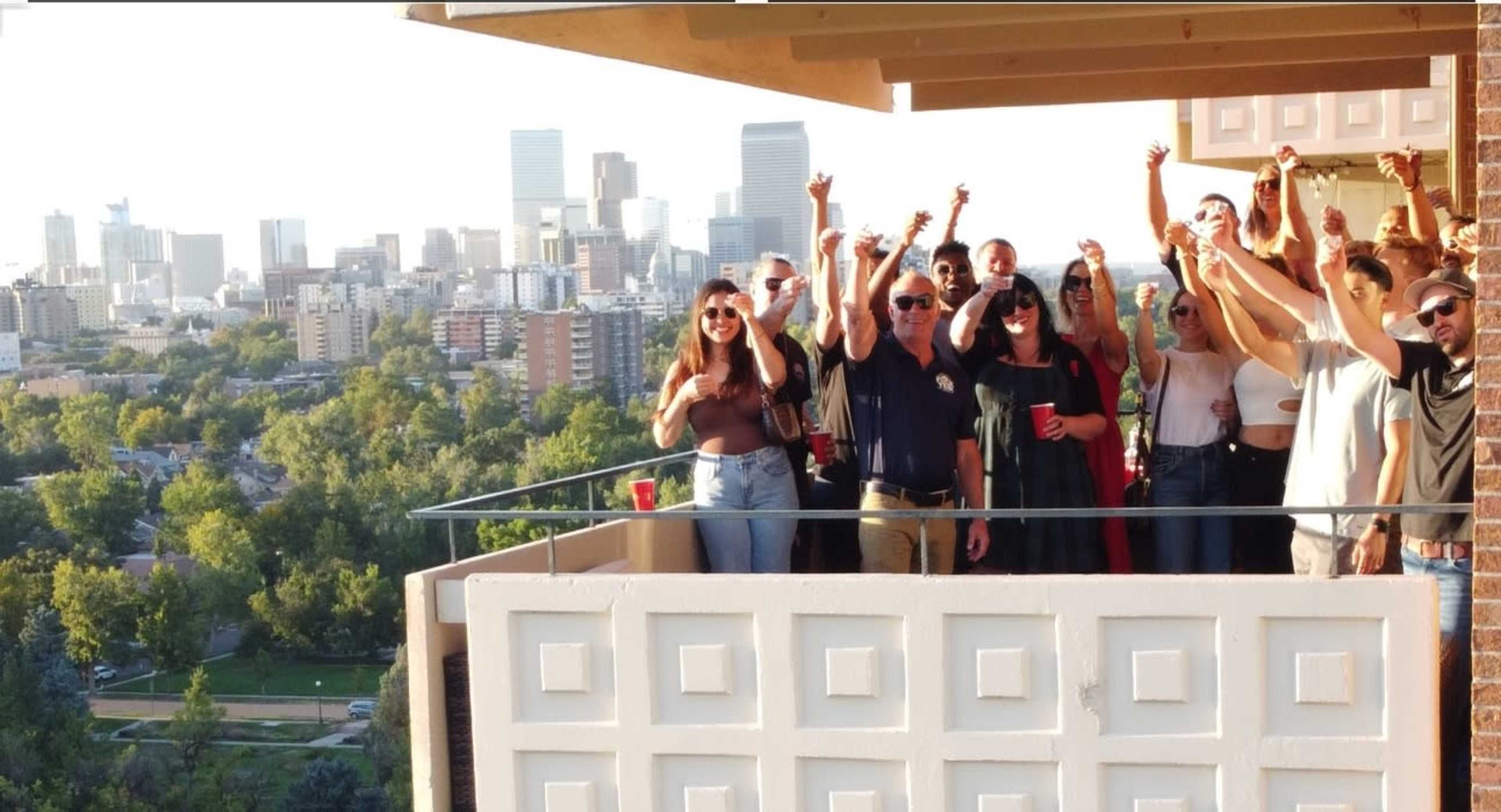 A group of people raises their arms in celebration on a balcony overlooking a city skyline.