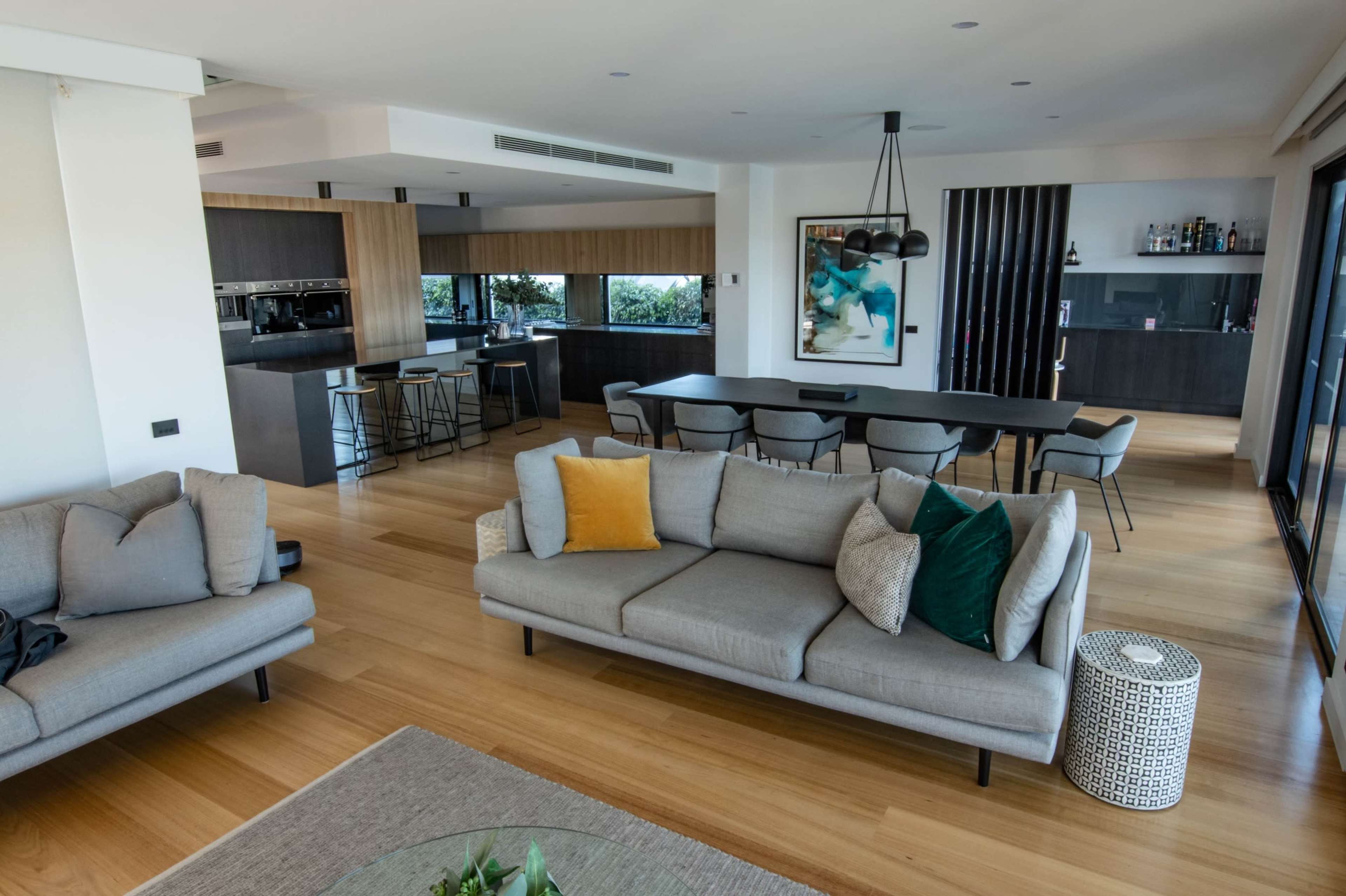 A modern living room with a gray sofa, a coffee table, and a dining area featuring a large table and contemporary chairs, leading to an open kitchen.
