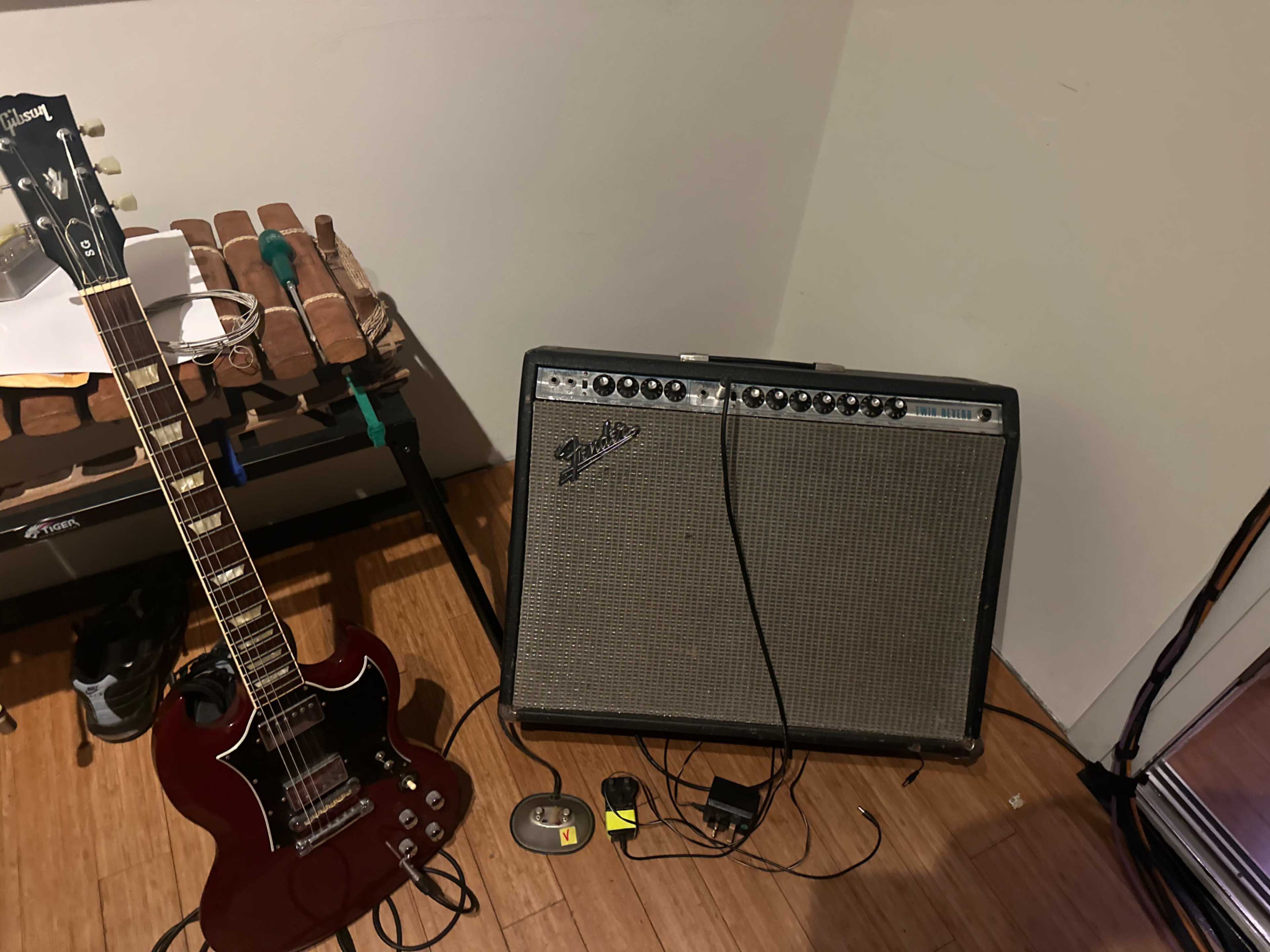 A red electric guitar leans against a music amplifier on a wooden floor, with additional equipment visible nearby.