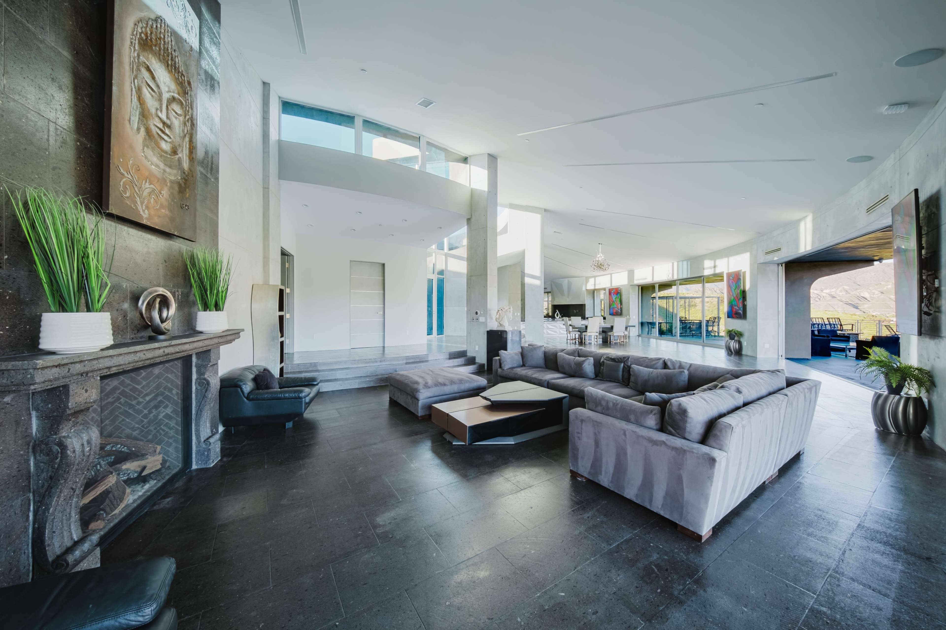 The image shows a spacious modern living room with large windows, a sectional sofa, and a stone fireplace, featuring minimalist decor and potted plants.