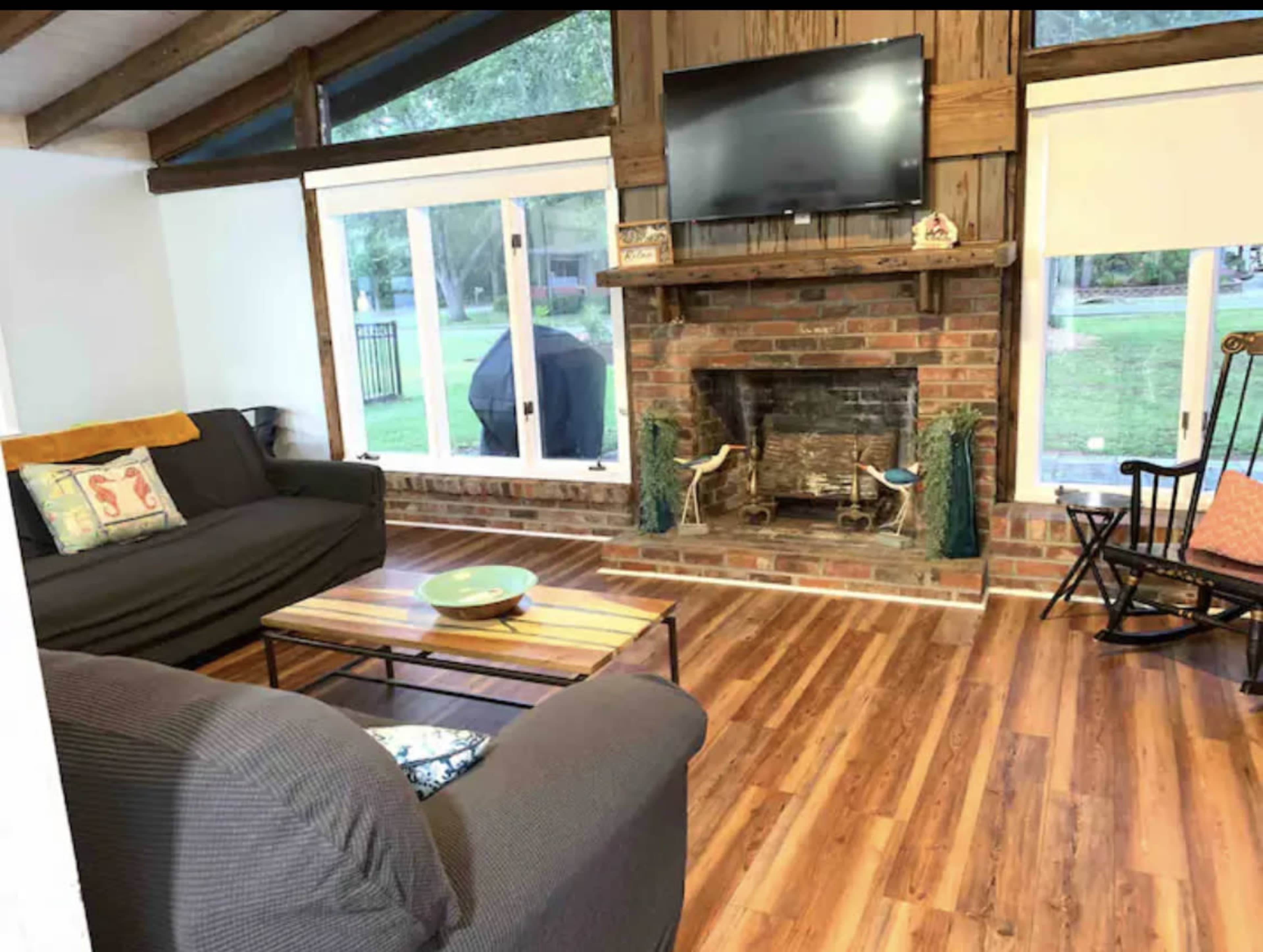 The image shows a living room with a brick fireplace, a television mounted above it, two sofas, a wooden coffee table, and large windows overlooking a yard.
