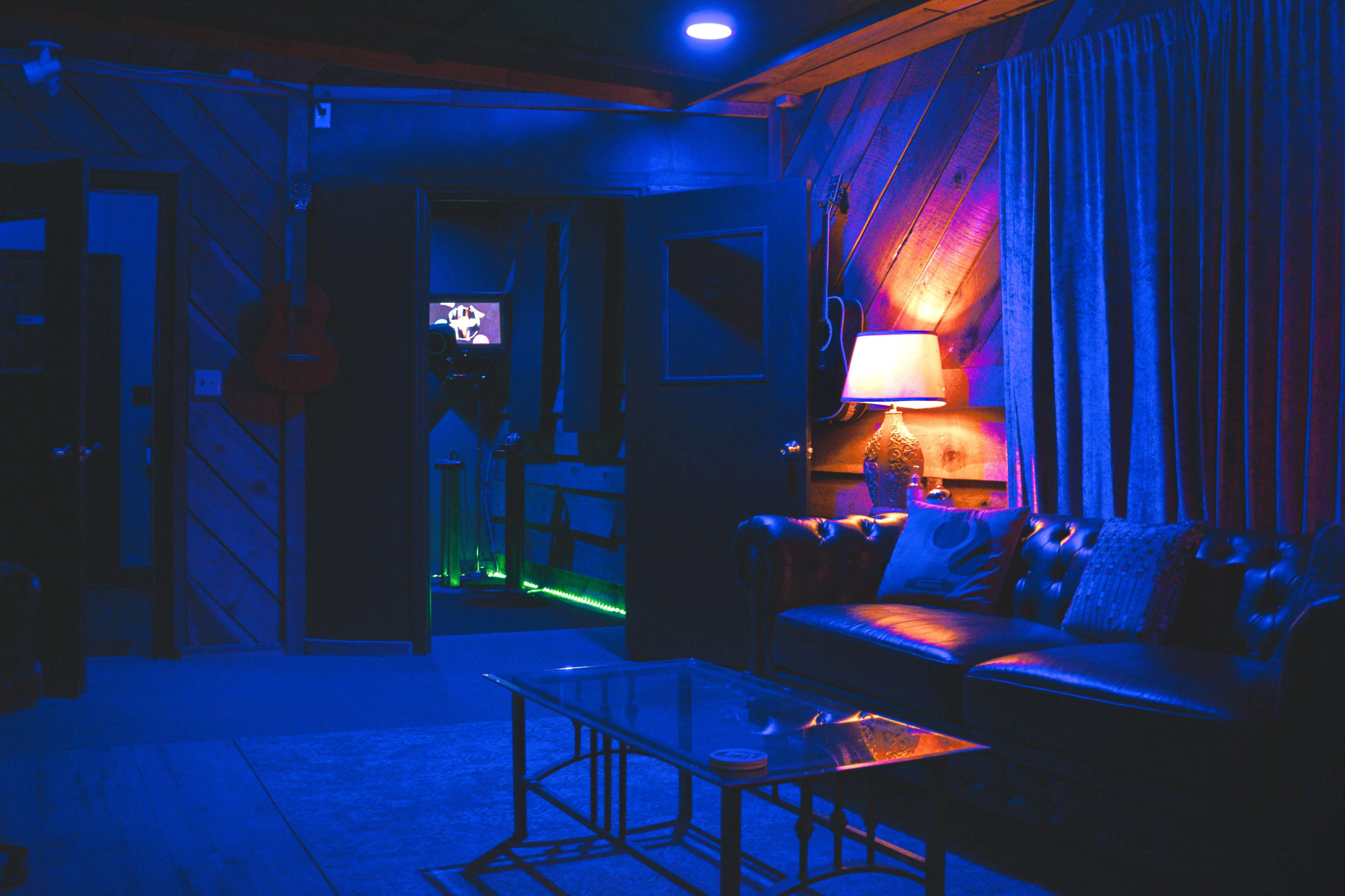 A dimly lit room features a brown leather couch, a coffee table, and doors leading to another area illuminated by colored lights.
